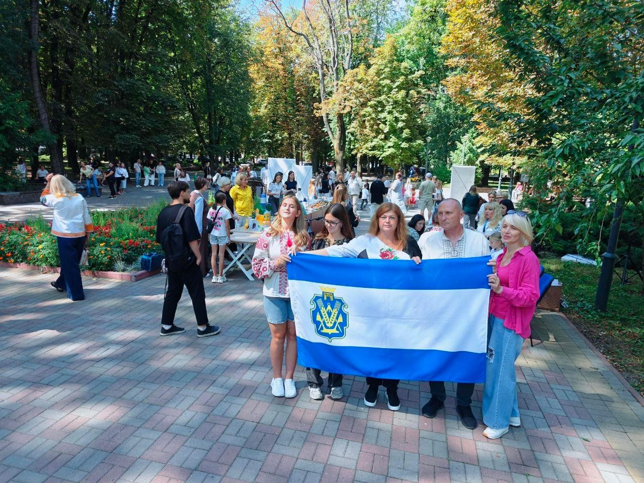 Начальник Костянтинівської СВА Олексій Кільдеров разом із фахівцями координаційного центру «Вільні разом» та молоддю позирують для фото на алеї міського парку під час молодіжного фестивалю «Покоління Незалежних» у Хмельницькому. Група людей тримає перед собою великий синьо-білий прапор із гербом міста Херсон. Серед присутніх — чоловік та дівчина у традиційних українських вишиванках. На задньому плані видно інших відвідувачів заходу та облаштовані просто неба виставкові столи на тлі зелених паркових дерев.