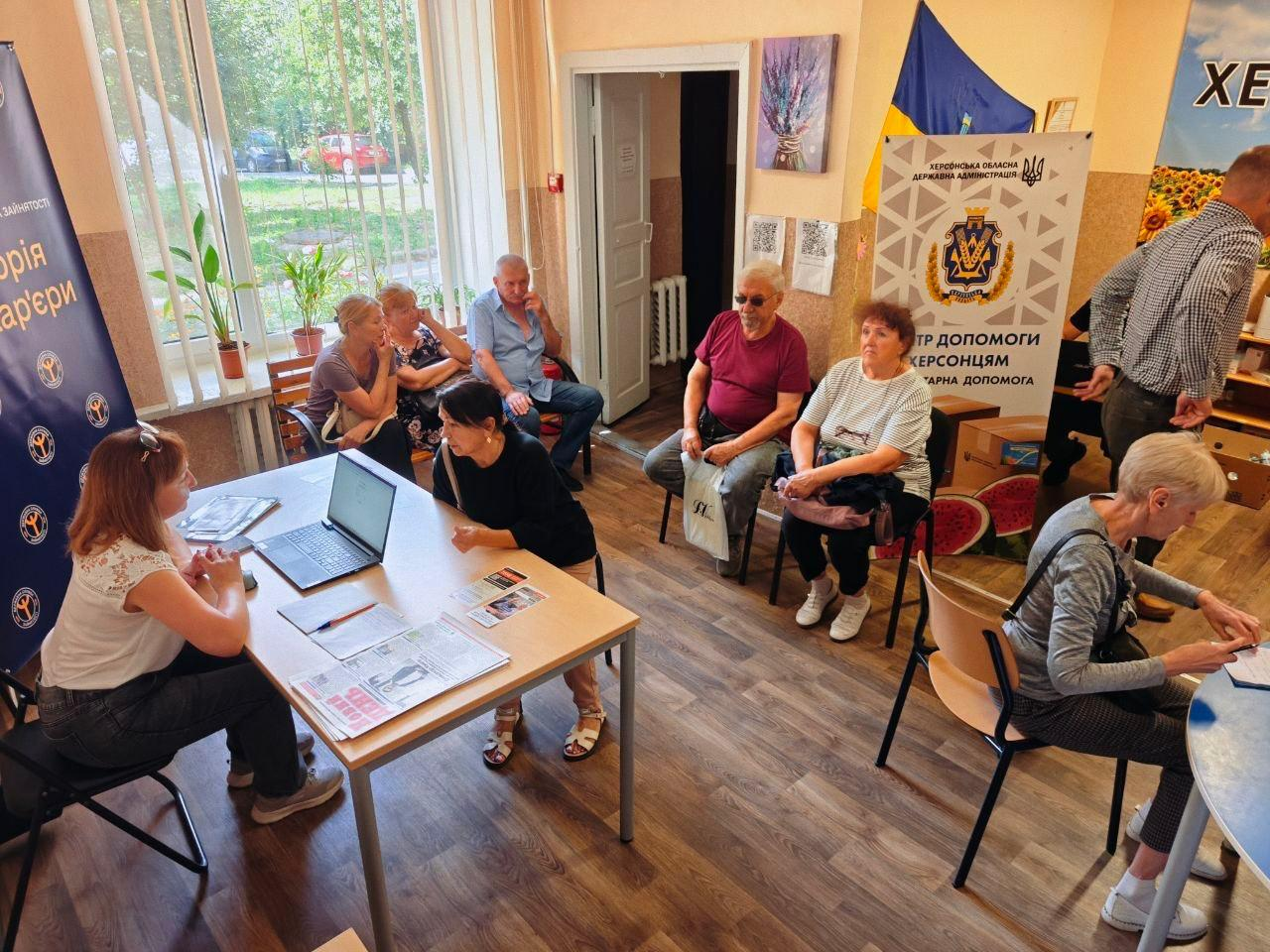Працівники ЦНАП Великолепетиської та Костянтинівської громад надають послуги та консультують переселенців у Хмельницькому координаційному центрі «Вільні разом». Ліворуч за робочим столом сидить фахівчиня перед відкритим ноутбуком і розкладеними бланками, спілкуючись із відвідувачкою. У залі на стільцях та лавках чекають своєї черги інші люди старшого віку. На задньому плані праворуч встановлено білий інформаційний банер Херсонської ОДА з гербом області та написом «Центр допомоги херсонцям. Гуманітарна допомога», а ліворуч — велике вікно, що виходить на зелену вулицю.