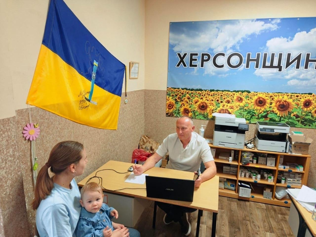 Начальник Костянтинівської СВА Олексій Кільдеров за робочим столом у Хмельницькому координаційному центрі «Вільні разом» консультує молоду жінку з маленькою дитиною на колінах. Перед керівником на столі стоїть відкритий ноутбук. Позаду нього на стіні розміщено великий фотобанер із написом «ХЕРСОНЩИНА» на тлі соняшникового поля та блакитного неба. Ліворуч на стіні закріплено державний прапор України з малюнком і підписами, а праворуч видно полиці з оргтехнікою та документами.