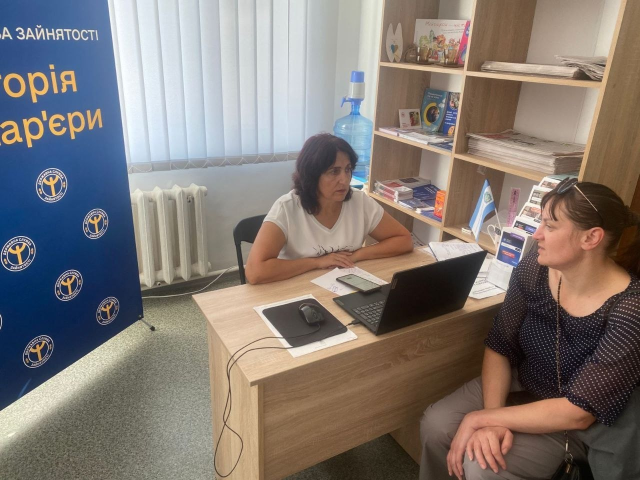 Фахівчиня Бериславської філії Херсонського обласного центру зайнятості консультує переселенку з Херсонщини в Житомирському координаційному центрі «Вільні разом». Дві жінки сидять за світлим дерев'яним столом. Працівниця у білій футболці сидить перед відкритим ноутбуком, а навпроти неї — відвідувачка у темній блузці в горошок. Ліворуч встановлено темно-синій банер служби зайнятості з її логотипами та помаранчевим гаслом «Траєкторія кар'єри». На задньому плані стоїть дерев'яний стелаж із друкованою продукцією. Супровідний текст зазначає, що в межами цієї всебічної підтримки ВПО консультації з питань працевлаштування отримали 8 осіб.
