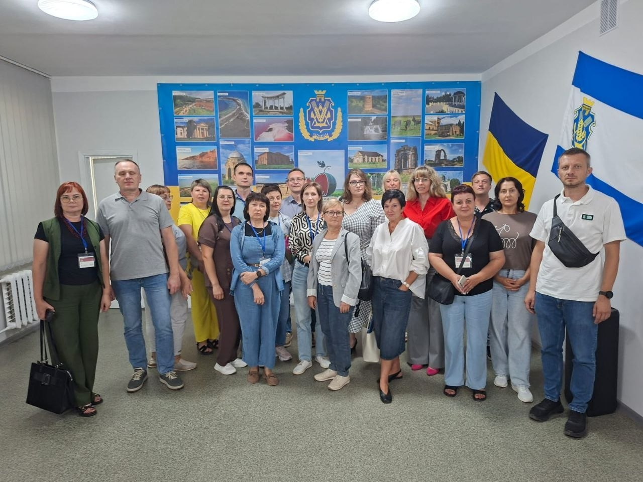Велика група переселенців із Херсонщини та працівників Житомирського координаційного центру «Вільні разом» позирує для спільного фото в офіційному кабінеті під час заходу всебічної підтримки внутрішньо переміщених осіб. Близько двадцяти людей різного віку стоять разом на тлі великого синього настінного стенда з фотоколажем пам'яток Херсонщини, її гербом та символічним малюнком кавуна. Праворуч на стіні розгорнуто великий синьо-білий прапор міста Херсон та державний прапор України. У декого з присутніх на шиї помітні сині брендовані стрічки з бейджами.