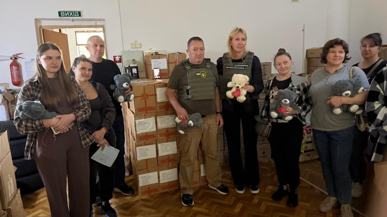 На фотографії зафіксовано зустріч начальника Чулаківської СВА Руслана Русаля та директорки благодійної організації Тетяни Грибачевої з жителями Херсонщини у транзитному центрі під час передачі гуманітарної допомоги від БО «БФ «ХУМЕІД». У центрі кадру стоять Руслан Русаль та Тетяна Грибачева, одягнені в однакові за кольором захисні бронежилети хакі, але з різними нашивками: у начальника СВА на грудях закріплено патч із гербом України, а у директорки фонду — білий текстовий напис. Тетяна Грибачева тримає в руках невелике біле плюшеве ведмежа з червоним серцем. Навколо керівників зібралася група місцевих жительок та волонтерів, які також тримають у руках подаровані м'які іграшки — сірих ведмедиків. Вони позирують у світлому приміщенні хабу на тлі високих стопок із картонними коробками, у яких доставлено одяг, солодощі, іграшки та канцелярське приладдя.