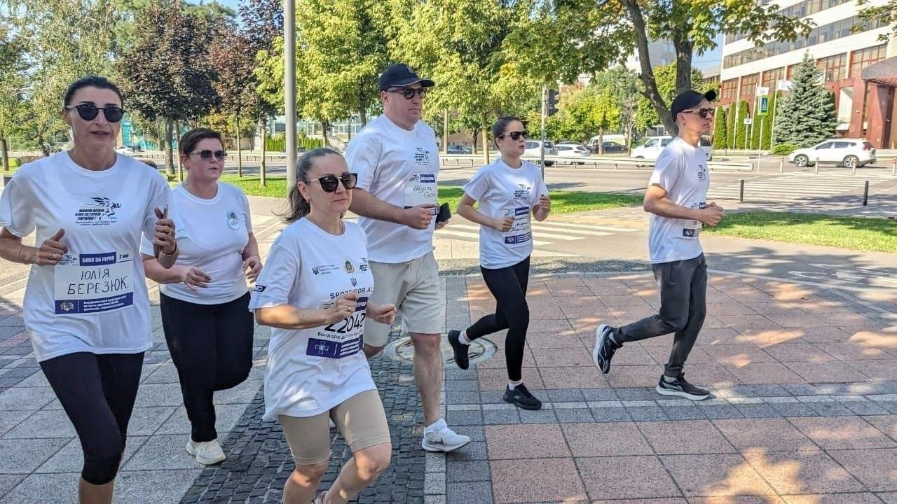 На фотографії зафіксовано участь херсонських переселенців та представників координаційного центру «Вільні разом» у щорічному патріотичному забігу «Шаную воїнів, біжу за Героїв України» у Дніпрі. Група із шести бігунів у сонцезахисних окулярах рухається брукованою доріжкою міського парку на тлі дерев та проїжджої частини. Усі учасники одягнені у фірмові білі спортивні футболки заходу із символікою забігу та закріпленими на грудях персональними номерами; зокрема, на передньому плані ліворуч біжить жінка з іменним номером на честь Юлії Березюк.