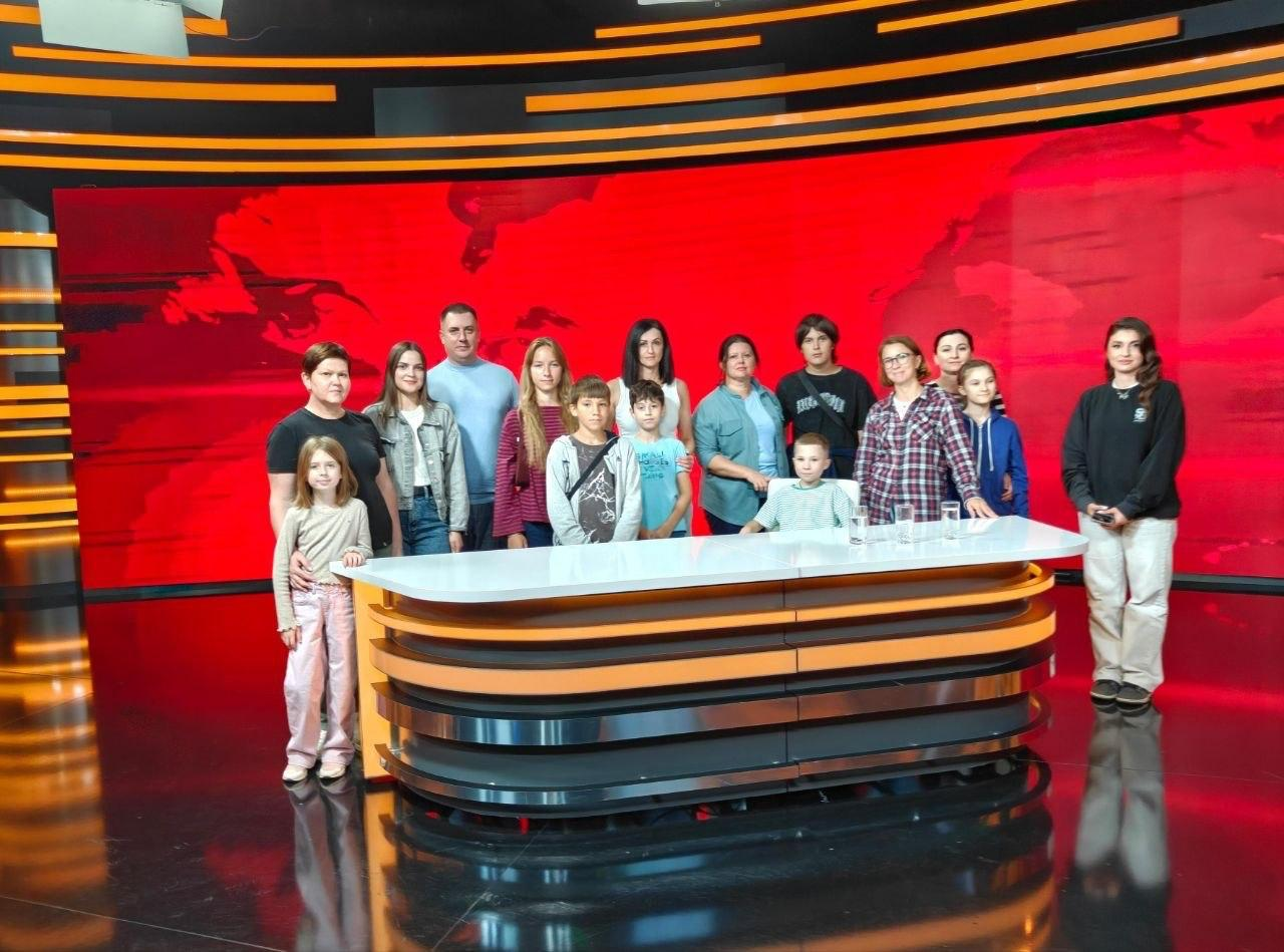 На фотографії зафіксовано екскурсію за лаштунками телеканалу «ДніпроTV», організовану для юних херсонських переселенців координаційним центром «Вільні разом» у Дніпрі. Група дітей та дорослих стоїть у два ряди навколо півкруглого сучасного студійного столу зі світлою глянцевою поверхнею та помаранчево-чорними декоративними смугами. Учасники екскурсії одягнені у повсякденний одяг і позирують у професійній телевізійній студії новин на тлі великого яскраво-червоного екрана-задника із зображенням карти світу, елементи якого віддзеркалюються у темній підлозі.