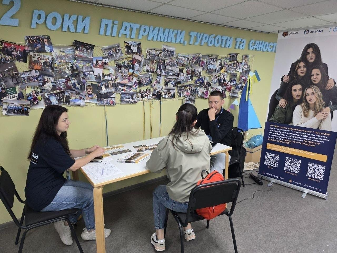 На фотографії зафіксовано процес надання індивідуальних юридичних консультацій внутрішньо переміщеним особам із Херсонщини від фахівців партнерських організацій у координаційному центрі «Вільні разом» у Дніпрі. За світлим дерев'яним столом, де розкладено інформаційні буклети, сидить представниця ГО «Art of Aid» у чорній фірмовій футболці та її колега-консультант, які спілкуються з дівчиною-відвідувачкою. Зустріч, під час якої 15 переселенців отримали фахову допомогу від БФ «Рокада», БФ «Право на захист» та ГО «Art of Aid», проходить на тлі стіни з великим написом «2 РОКИ ПІДТРИМКИ, ТУРБОТИ ТА ЄДНОСТІ», яскравим фотоколажем та інформаційним банером проєкту із QR-кодами організації.