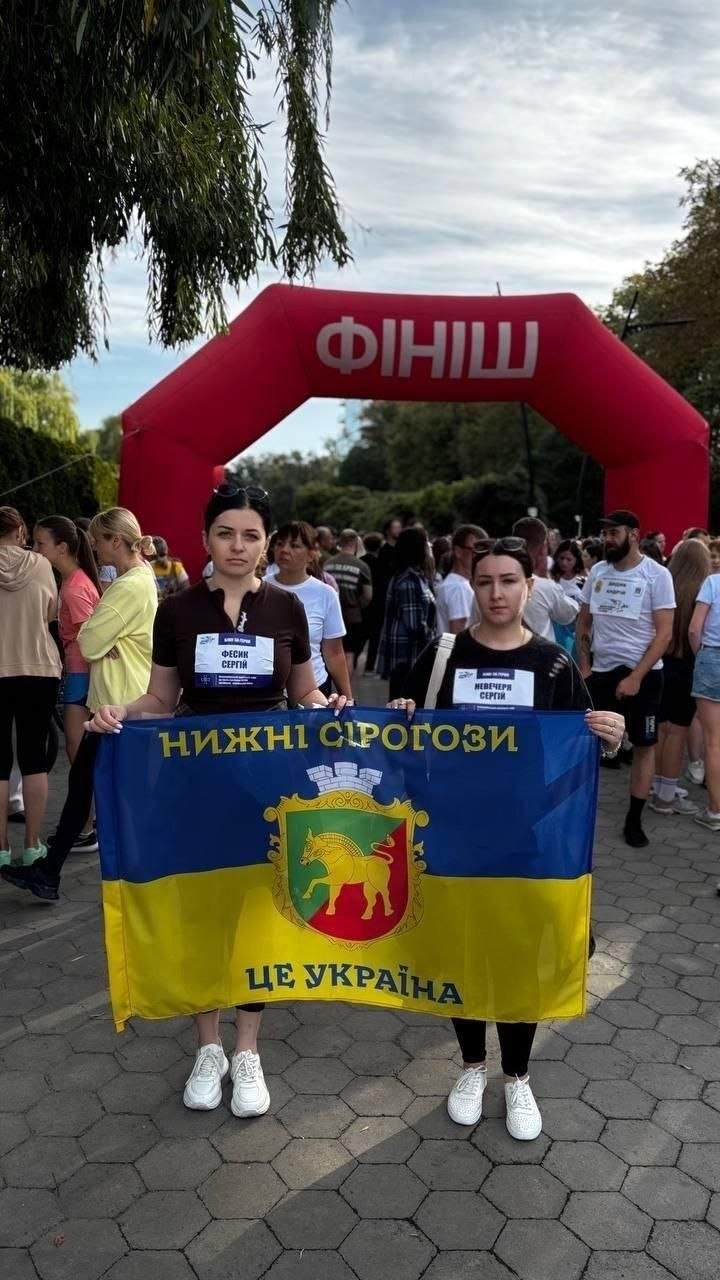 Представниці Нижньосірогозької громади та переселенці з Херсонщини беруть участь у традиційному щорічному забігу «Шаную воїнів, біжу за героїв України» до Дня пам’яті захисників України. Дві дівчини стоять на першому плані та тримають розгорнутий синьо-жовтий прапор із гербом селища Нижні Сірогози, написом «Нижні Сірогози» зверху та «Це Україна» знизу. Учасниці вдягнені у темні футболки й білі кросівки, а на їхніх грудях прикріплені стартові номери забігу імені полеглих воїнів-земляків (на лівому номері написано «Фесик Сергій»). Позаду дівчат на брукованій алеї стоїть великий натовп інших бігунів, а на задньому плані височіє велика червона надувна арка з білим написом «Фініш», оточена зеленими деревами.