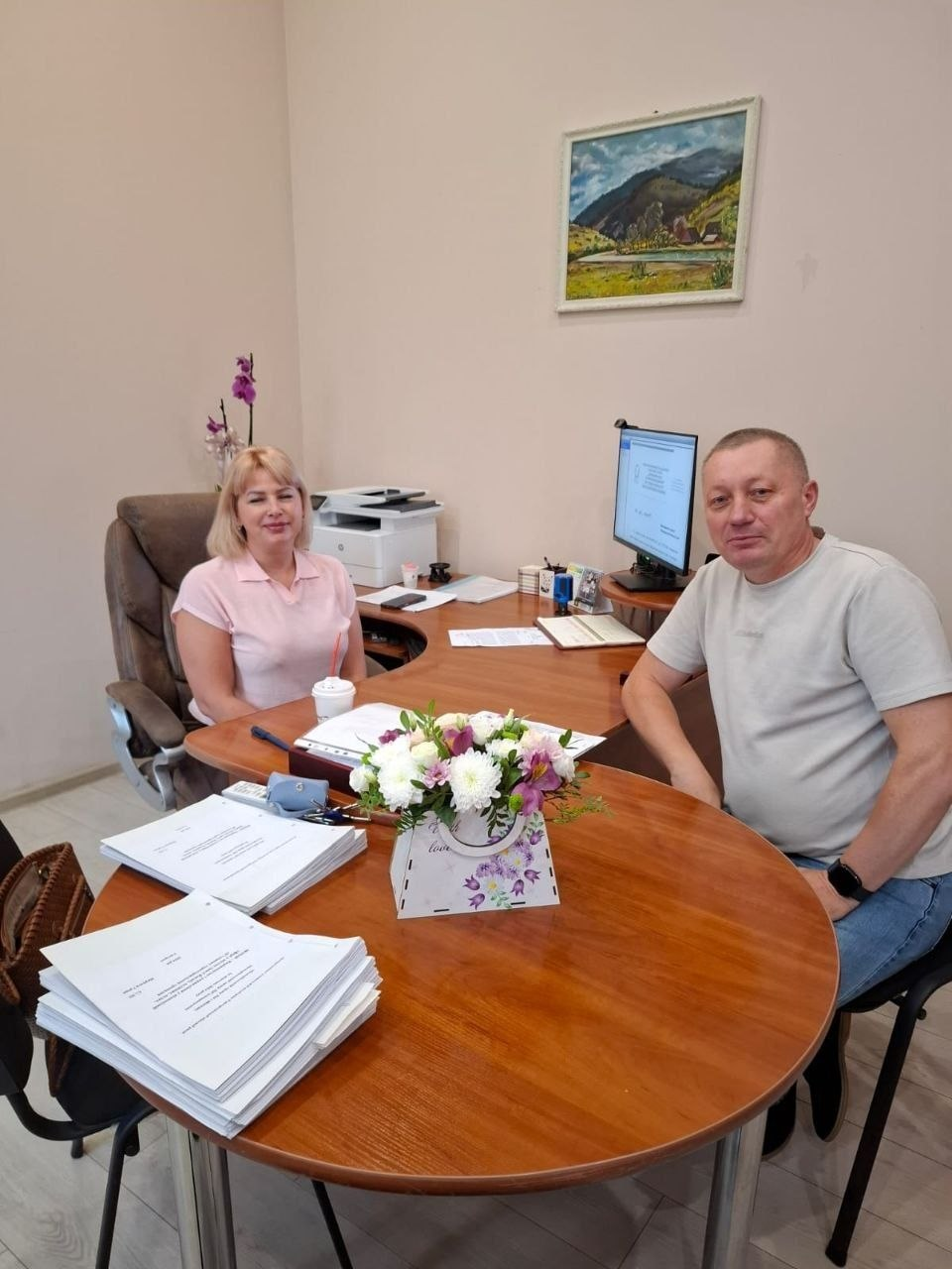Начальник Нижньосірогозької СВА Віталій Кривчик проводить особистий прийом громадян в ужгородському координаційному центрі «Вільні разом». Чоловік у світлій футболці та джинсах сидить праворуч за великим кутовим дерев'яним столом. Навпроти нього, у м'якому офісному кріслі ліворуч, сидить відвідувачка — блондинка у світло-рожевій футболці-поло. На круглому краї столу перед ними лежать дві великі стопки офіційних документів А4, а між ними стоїть квіткова композиція з білих та бузкових хризантем у коробці-кашпо. Робоче місце обладнане увімкненим монітором комп'ютера, канцелярським приладдям, телефоном та білим БФП на задньому плані. Стіни кабінету світлі, а над комп'ютером висить картина з гірським краєвидом.