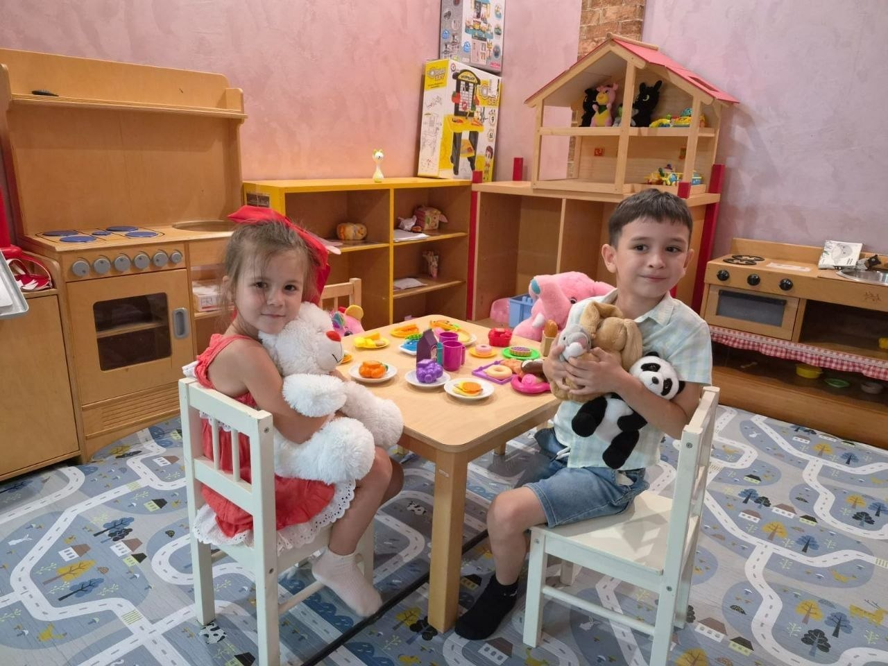 Маленькі відвідувачі дитячого простору в ужгородському координаційному центрі «Вільні разом» граються в ігровій кімнаті. Двоє дітей — дівчинка та хлопчик — сидять навпроти одне одного за маленьким світлим столиком на білих дерев'яних стільчиках. Ліворуч посміхається дівчинка в рожевій сукні з великим червоним бантом у волоссі, обіймаючи білого плюшевого ведмедика. Праворуч сидить хлопчик у світлій сорочці та шортах, тримаючи в руках іграшкову панду та зайчика. На столі між ними розставлено різнокольоровий дитячий посуд для чаювання. На підлозі лежить сірий килим із принтом автомобільних доріг, а на задньому плані облаштовано дитячу зону з іграшковою дерев'яною кухнею, стелажами та великим ляльковим будиночком.