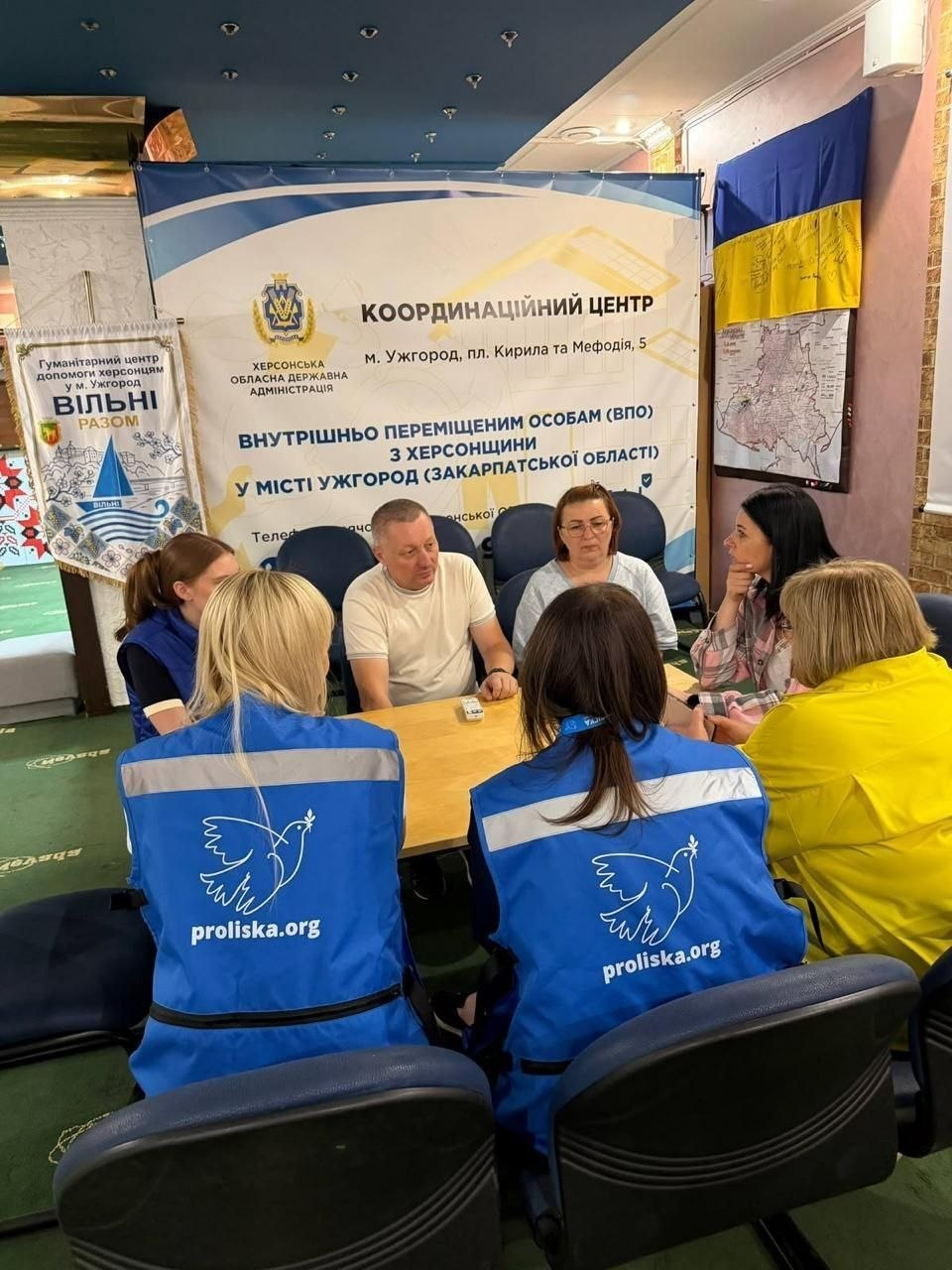 Начальник Нижньосірогозької селищної військової адміністрації Віталій Кривчик (сидить по центру в білій футболці) бере участь у зустрічі в ужгородському координаційному центрі «Вільні разом». Разом із ним за великим столом сидять відвідувачі та представниці гуманітарної місії «Проліска». На першому плані спиною до камери сидять дві жінки-фахівчині у яскраво-синіх жилетах місії з білим логотипом птаха та адресою сайту proliska.org, які проводять засідання групи психологічної підтримки та надають консультації. На задньому плані розміщено великий офіційний банер «Координаційний центр внутрішньо переміщеним особам (ВПО) з Херсонщини у місті Ужгород» із гербом Херсонської ОДА. На стіні праворуч висить український прапор та карта, а ліворуч стоїть банер гуманітарного центру «Вільні разом».