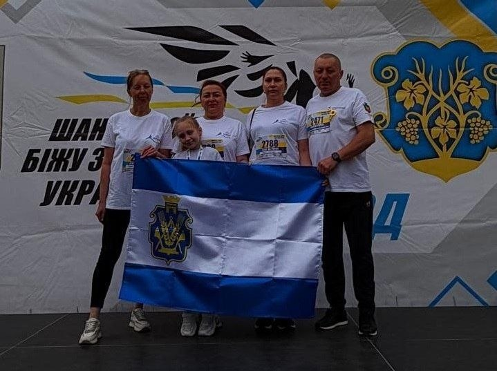 Начальник Нижньосірогозької селищної військової адміністрації Віталій Кривчик разом із представниками громади та переселенцями з Херсонщини бере участь у патріотичному забігу «Шаную воїнів, біжу за героїв України» в межах церемонії вшанування пам’яті полеглих захисників. На сцені стоять п'ятеро учасників акції: четверо дорослих та одна маленька дівчинка. Усі вони вдягнені у білі фірмові футболки заходу зі стартовими номерами учасників. Група тримає перед собою розгорнутий синьо-біло-синій прапор із гербом Херсонської області. Вони позирують на тлі великого світлого тематичного банера з гербом, національним орнаментом та офіційним гаслом забігу «ШАНУЮ ВОЇНІВ, БІЖУ ЗА ГЕРОЇВ УКРАЇНИ».