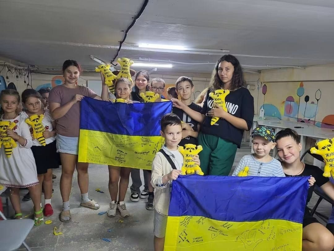Групова фотографія дітей та підлітків під час патріотичного виховного заняття, організованого фахівцями Центру соціальної підтримки дітей та сімей Херсонської обласної ради. Учасники посміхаються та тримають два великі державні прапори України — синьо-жовті полотнища, на жовтій частині яких діти залишили численні рукописні побажання, автографи та патріотичні малюнки. Багато хто з дітей тримає в руках однакові символічні сувеніри — м'які жовті іграшки у формі ведмедиків із написами «Живи, Херсон! Живи!!!». Зустріч проходить у приміщенні з низькою стелею, стіни якого прикрашені яскравими дитячими малюнками кулястих дерев та повітряних кульок.