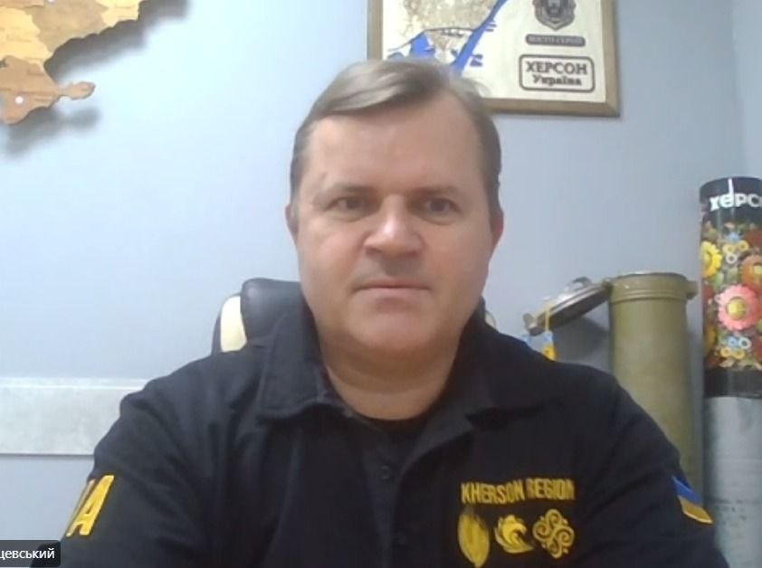 Скриншот з онлайн-трансляції засідання Гуманітарного штабу при Херсонській ОВА під керівництвом заступника голови Херсонської ОДА Володимира Клюцевського. Посадовець сидить в офісному кріслі та дивиться в камеру під час виступу. На ньому чорний формений одяг, на грудях якого вишито жовтий напис «KHERSON REGION» та три золотаві емблеми-символи, а на лівому рукаві розміщено прапор України. У лівому нижньому кутку екрана видно плашку з частиною його прізвища «...цевський». Зустріч проходить на тлі світло-сірої стіни кабінету, де ліворуч закріплено дерев'яну карту України, по центру висить тематична картина в рамці з написом «ХЕРСОН Україна», а праворуч стоять військовий тубус від артилерійського снаряда та патріотично розписаний квітковими візерунками гільзовий елемент.
