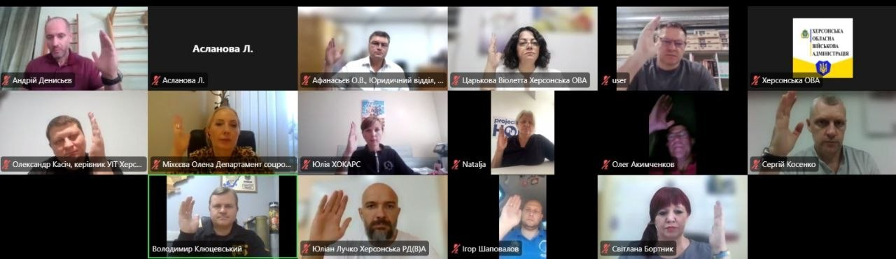 Скриншот екрана під час засідання Гуманітарного штабу при Херсонській ОВА, яке проводиться в режимі онлайн-відеоконференції. Монітор розділений на сітку з 15 вікон, у яких присутні учасники заходу — посадовці та керівники профільних департаментів та відомств. Більшість членів штабу одночасно тримають підняту вгору руку, демонструючи одностайне голосування за рішення про розподіл допомоги громадам. Усі віконця мають офіційні підписи, серед яких видно імена: Андрій Денисьєв, Афанасьєв О.В., Царькова Віолетта, Олександр Касіч, Міхєєва Олена, Юлія ХОКАРС, Олег Акимченков, Сергій Косенко, Юліан Лучко, Ігор Шаповалов, Світлана Бортник та заступник голови Херсонської ОДА Володимир Клюцевський (вікно якого підсвічене зеленим контуром). В одному з вікон відображається лише чорний екран із підписом «Асланова Л.», а у правому верхньому кутку розміщено офіційну емблему Херсонської обласної військової адміністрації.