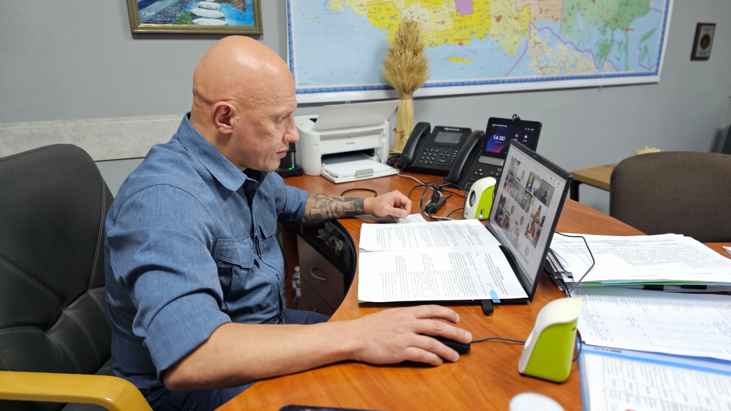 Заступник начальника Херсонської ОВА Євгеній Ігнатенко проводить чергове засідання обласної комісії з питань надання матеріальної допомоги жителям області. Чоловік міцної статури з поголеною головою та татуюванням на лівій руці сидить у великому чорному шкіряному кріслі за великим дерев'яним кутовим столом. Він вдягнений у синю сорочку з коротким рукавом і працює за ноутбуком, на екрані якого відображається онлайн-відеоконференція з учасниками засідання. Перед ним на столі лежать роздруковані заяви громадян та офіційні документи формату А4. Робоче місце обладнане білим принтером, стаціонарним телефоном, мережевими пристроями та канцелярією в біло-зелених підставках. На сірій стіні кабінету позаду посадовця висить велика настінна карта Херсонської області, поруч із якою на тумбі стоїть декоративний сніп пшениці.