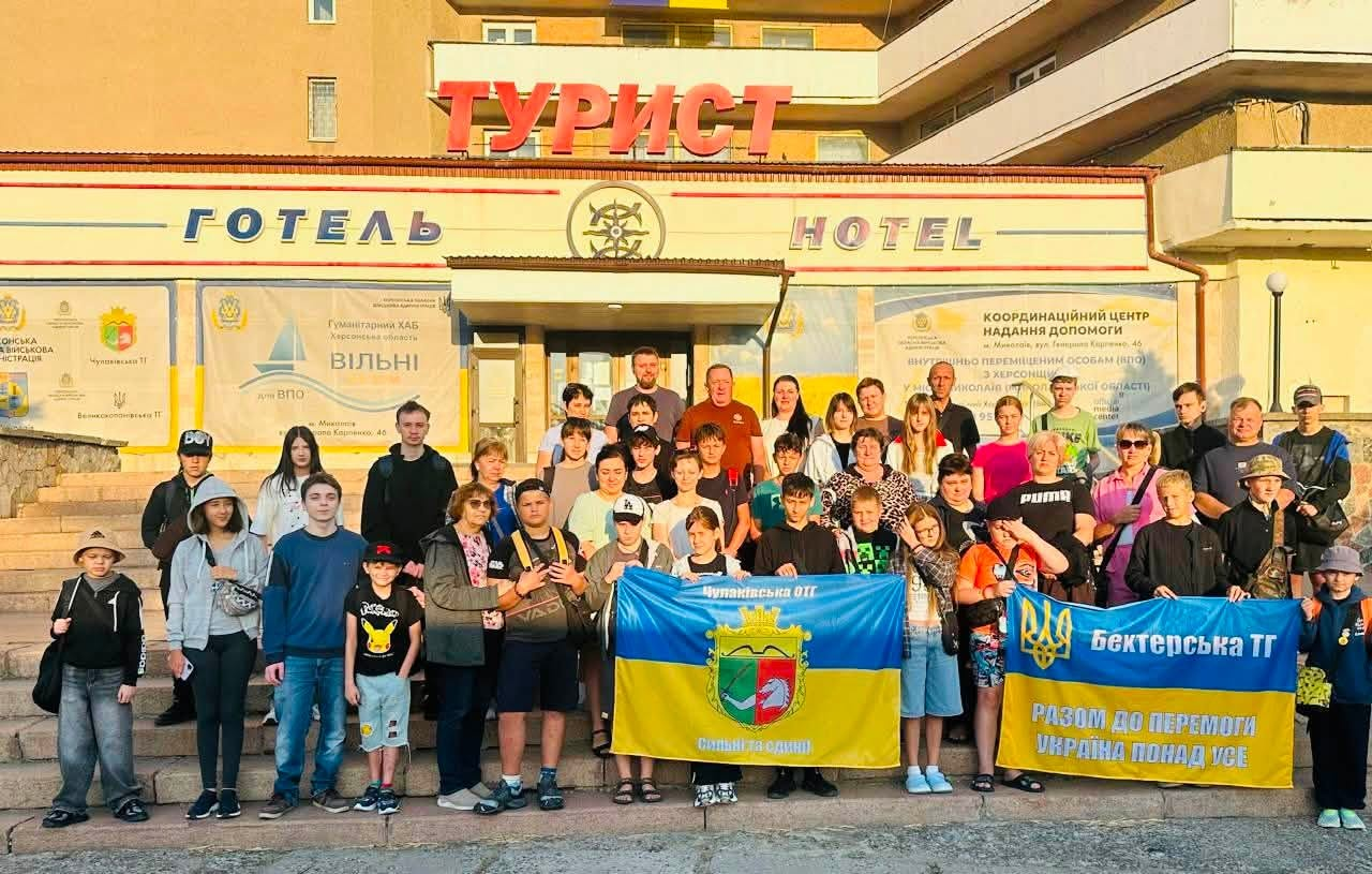 Група з 30 дітей-переселенців із Херсонщини, їхніх батьків та організаторів поїздки стоїть на широких кам'яних сходах перед входом до готелю «Турист» у Миколаєві, де діє координаційний хаб. На першому плані учасники тримають два великі прапори громад Скадовського району. Ліворуч — синьо-жовтий прапор із гербом та написом «Чулаківська ОТГ. Сильні та єдині». Праворуч — синій прапор із тризубом, написом «Бехтерська ТГ» та жовтою смугою знизу з гаслом «РАЗОМ ДО ПЕРЕМОГИ. УКРАЇНА ПОНАД УСЕ». Серед присутніх у центрі натовпу — начальник Чулаківської СВА Руслан Русаль та начальник Бехтерської СВА Олексій Баткалов. Над входом у будівлю встановлено великі червоні літери «ТУРИСТ» та вивіски «ГОТЕЛЬ / HOTEL». По боках від дверей розміщено інформаційні банери Координаційного центру надання допомоги ВПО з Херсонської області у Миколаєві.