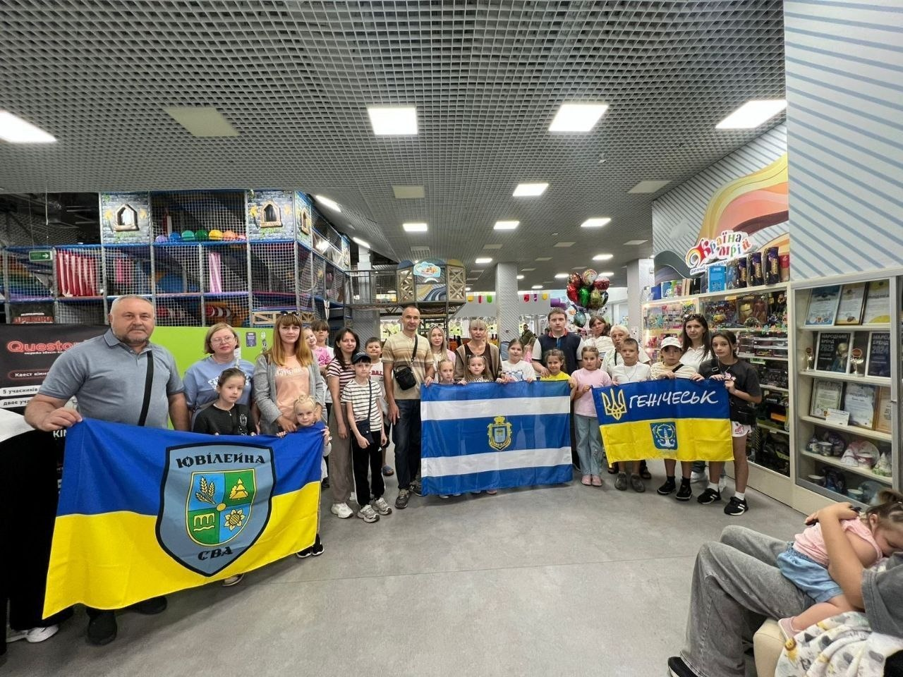 Група переселенців із Херсонщини — дітей та дорослих — позує для спільного фото під час заходу, організованого координаційним центром «Вільні разом» у Вінниці. Учасники зустрічі розгорнули у великому дитячому розважальному комплексі три офіційні прапори громад: синьо-жовтий прапор Ювілейної сільської військової адміністрації (СВА), біло-синій прапор Херсонської області та державний прапор із гербом і написом «Генічеськ». На задньому плані приміщення видно ігровий лабіринт, вітрини з іграшками та стелажі з грамотами.