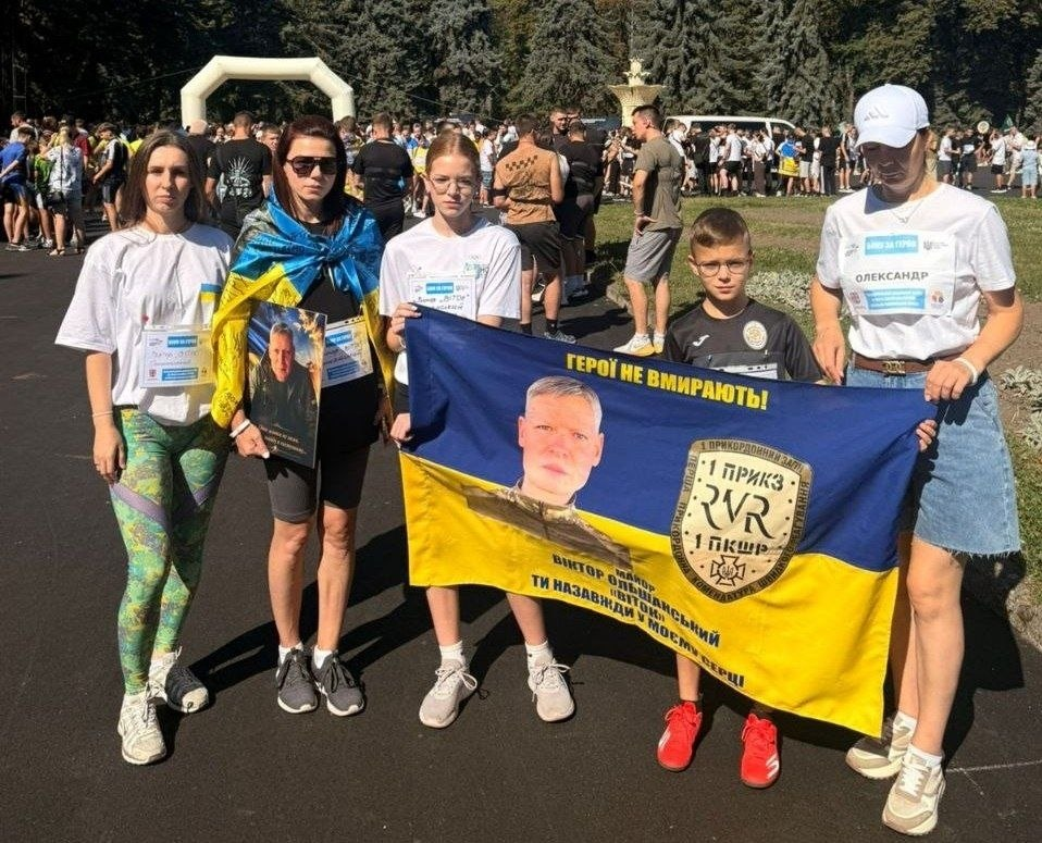 Переселенці з Херсонщини та представники координаційного центру «Вільні разом» у Вінниці беруть участь у щорічному забігу «Шаную воїнів, біжу за Героїв України». Група учасників тримає розгорнутий державний прапор із портретом майора Віктора Ольшанського, шевроном прикордонного загону та написом «Герої не вмирають!». Одна з учасниць ліворуч тримає меморіальний портрет іншого полеглого захисника, а на футболках бігунів прикріплені стартові номери забігу імені героїв-земляків. Акція проходить на асфальтованій парковій доріжці на тлі натовпу людей та білої стартової арки.