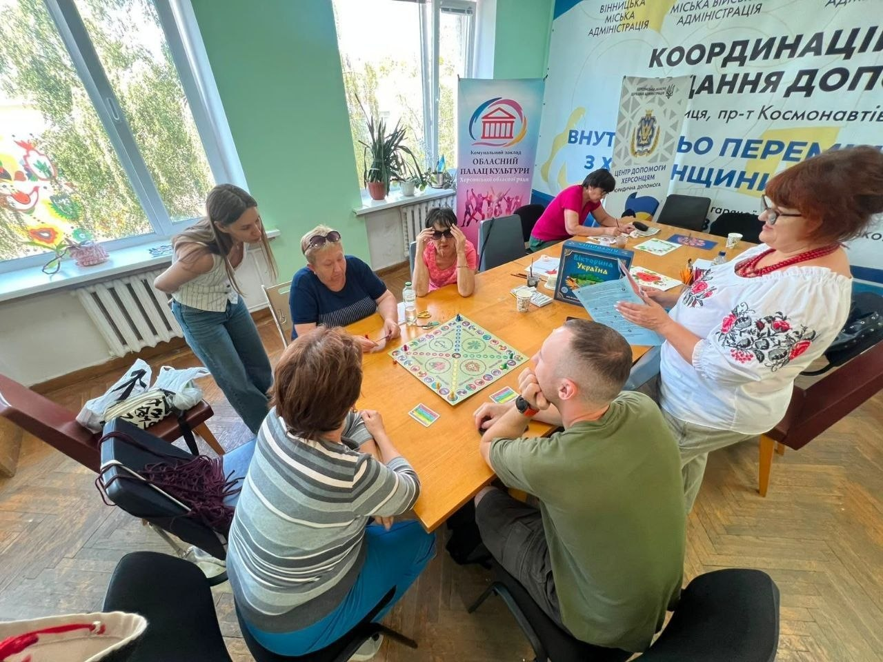 Група людей у світлій кімнаті координаційного центру «Вільні разом» у Вінниці грає в патріотичну настільну гру-вікторину «Україна» за великим дерев'яним столом. На передньому плані спиною до камери сидить військовослужбовець у камуфляжній футболці, який бере участь у грі. Жінка в білій вишиванці праворуч стоїть і демонструє учасникам ігрові картки та коробку вікторини. На стінах позаду розміщені банери з написами «Вінницька міська адміністрація» та інформаційні стенди центру допомоги внутрішньо переміщеним особам (ВПО) з Херсонщини.