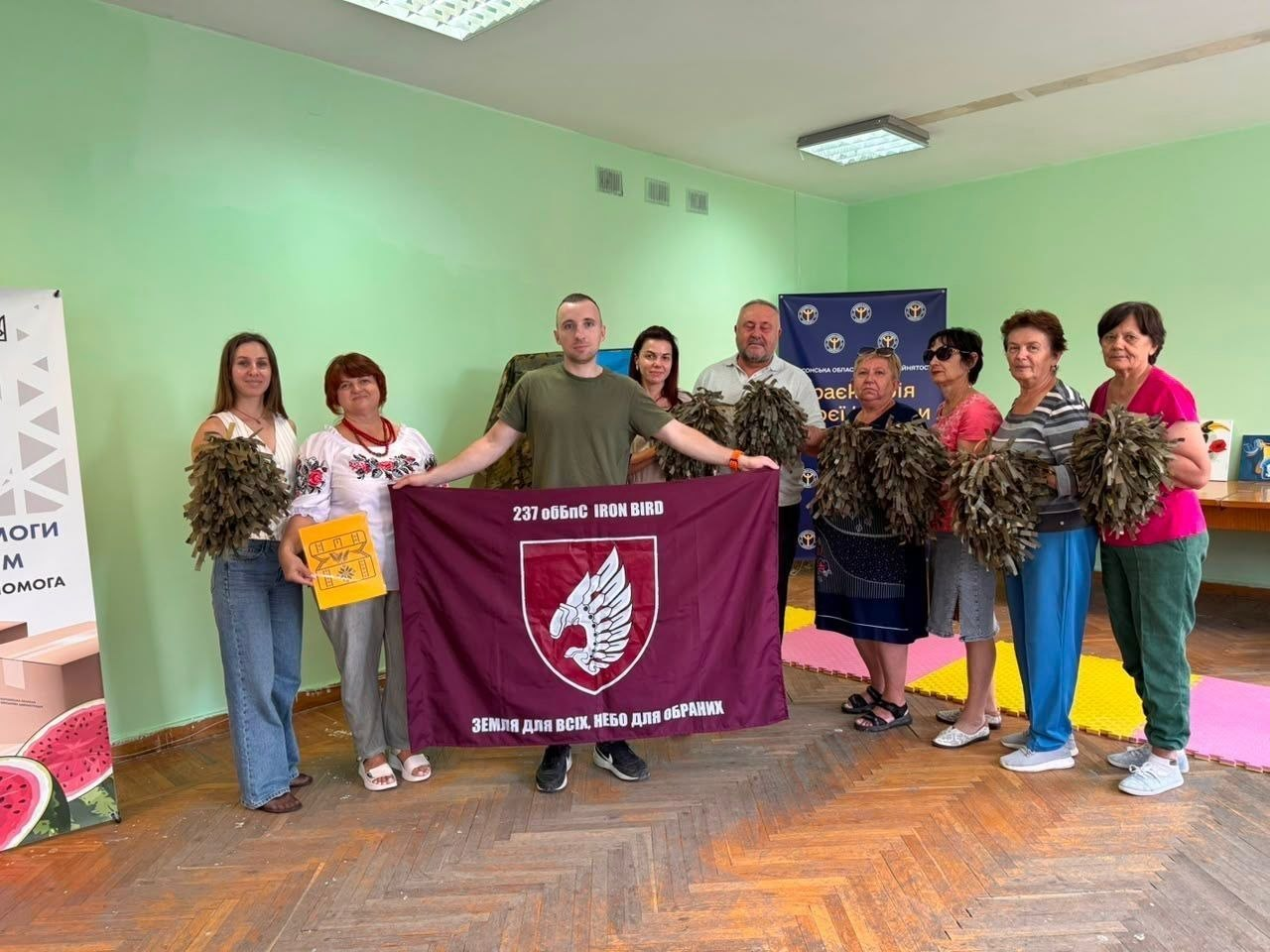 Переселенці з Херсонщини та організатори координаційного центру «Вільні разом» у Вінниці позують для фото з військовослужбовцем Сил оборони після передачі допомоги. Учасники зустрічі тримають у руках виготовлені для українських захисників та захисниць маскувальні нашоломники (елементи «кікімори»). По центру військовий у футболці хакі та жінки розгорнули темно-бордовий прапор 237-го окремого батальйону безпілотних систем «IRON BIRD» з білим крилатим гербом та гаслом «Земля для всіх, небо для обраних». На тлі світло-зелених стін зали також видно жовту папку та офіційний синій банер Херсонської обласної організації.