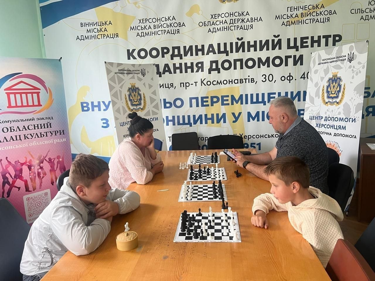 Двоє дітей та двоє дорослих беруть участь у турнірі з шахів та шашок для переселенців із Херсонщини в координаційному центрі «Вільні разом» у Вінниці. Вони сидять навпроти одне одного за довгим дерев'яним столом, на якому розставлені дві шахівниці з фігурами, а чоловік праворуч стежить за часом на мобільному телефоні. На задньому плані кабінету встановлено великий білий банер Координаційного центру надання допомоги ВПО з Херсонщини у місті Вінниця, а також інформаційні стенди Обласного палацу культури та Центру допомоги херсонцям.