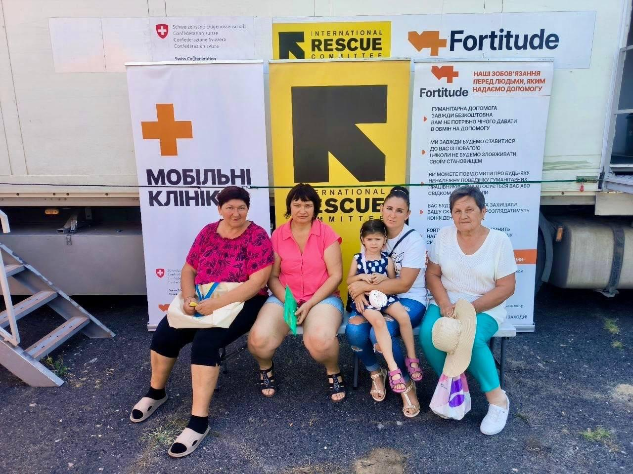Пацієнтки з дітьми чекають на прийом лікарів мобільної клініки БО «Благодійний фонд "Фортітьюд"» (Fortitude) у Бериславському районі. Троє жінок та дівчинка в літньому одязі сидять на лавці на тлі офіційних інформаційних банерів. Ліворуч розміщено банер із помаранчевим хрестом та написом «МОБІЛЬНІ КЛІНІКИ», по центру — великий жовтий стенд організації «International Rescue Committee» (IRC), а праворуч — плакат фонду «Fortitude» з інформацією про правила безкоштовного надання гуманітарної та медичної допомоги.
