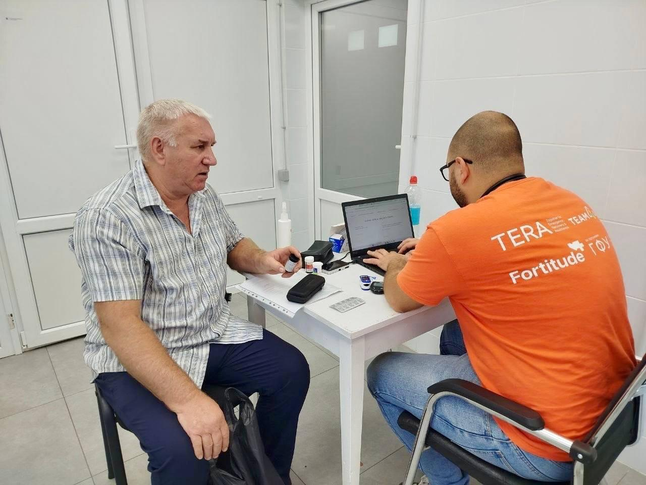 Лікар-терапевт команди «TERA TEAM» мобільної клініки «Fortitude» консультує пацієнта в Бериславському районі. Медик в окулярах та фірмовій помаранчевій футболці сидить спиною до камери та вносить дані в ноутбук. Чоловік у смугастій сорочці сидить навпроти за білим столом і тримає в руці баночку з безкоштовно отриманими ліками. На столі між ними лежать медичні прилади, пульсоксиметр, блістери з таблетками та антисептик. Зустріч проходить у чистому світлому приміщенні з металопластиковими дверима.