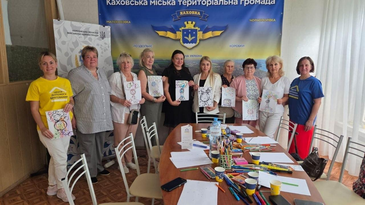 Учасниці сесії психологічного розвантаження з нейрографіки, організованої координаційним центром «Вільні разом» у Києві для переселенців із Херсонщини. Група жінок стоїть у кабінеті навколо великого столу та демонструє свої готові кольорові малюнки, створені під час арттерапії. На столі розкладено кольорові олівці, маркери та папір для малювання. На задньому плані височіє великий офіційний банер «Каховська міська територіальна громада» з гербом міста Каховка, а ліворуч встановлено інформаційний стенд Херсонської обласної військової адміністрації.