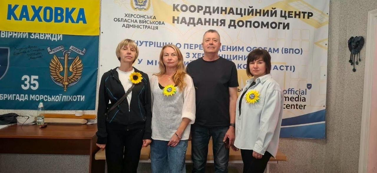 Команда координаційного центру «Вільні разом» у Києві та переселенці з Херсонщини беруть участь у меморіальному заході «Імена, які не можна забути. Соняхи пам’яті». Троє жінок та чоловік стоять у шеренгу, прикріпивши до одягу символічні жовті соняшники на знак вдячності полеглим воїнам. Вони позирують на тлі великого банера Координаційного центру надання допомоги ВПО з Херсонщини від Херсонської ОВА та офіційного медіацентра. Ліворуч на стіні кабінету розміщено синьо-жовтий стяг 35-ї окремой бригади морської піхоти з написом «КАХОВКА. ВІРНІ ЗАВЖДИ» та військовими автографами.