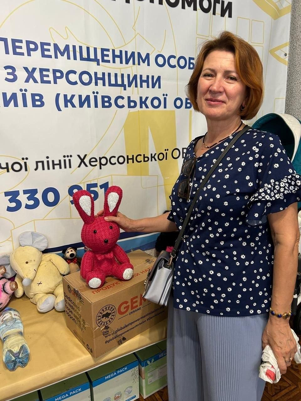 Жінка-переселенка з Херсонщини отримує продуктові набори та допомогу від координаційного центру «Вільні разом» у Києві. Вона посміхається і позирує на тлі великого офіційного банера Координаційного центру надання допомоги ВПО з Херсонщини. Жінка вдягнена у синю блузу в квіточку та спирається рукою на велику коробку з продуктовим набором від партнерів «GEM», на якій сидить яскраво-рожева в'язана іграшка-заєць. Поруч розкладено інші м'які іграшки, бутлі з питною водою та коробки з гуманітарною допомогою.