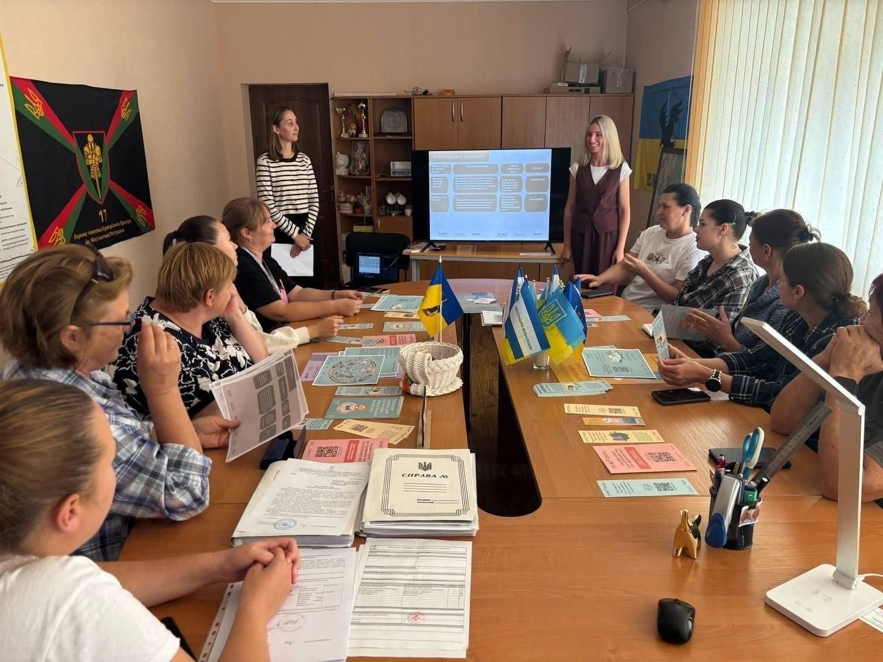 Учасники Дня спільнодії «Ти як? - Маршрут підтримки кожного в громаді» ознайомлюються з новими путівниками за великим робочим столом. На екрані в кінці кімнати демонструється презентація з блоками алгоритму скерування та перенаправлення пацієнтів, біля якого стоять координаторки заходу. Фахівці соціальної сфери, освіти та охорони здоров'я Борозенської громади сидять по обидва боки столу та вивчають розкладені друковані примірники «Маршрутів послуг з ментального здоров’я». На передньому плані видно офіційні папки з документами, а по центру столу — прапори України. Ліворуч на стіні розміщено військовий прапор 17-го окремого зенітного ракетного дивізіону.