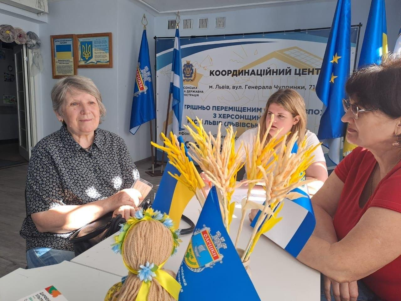 У приміщенні координаційного центру «Вільні разом» у місті Львів за столом проходять заходи інтерактивної лекції «Сакральні символи-обереги Херсонщини як відображення національної ідентичності українського народу». Ліворуч на фото — літня жінка-переселенка в темній блузі уважно слухає розповідь. По центру сидить молода фахівчиня хабу, яка проводить лекцію, а праворуч у червоній футболці — ще одна слухачка. На першому плані на білому столі стоїть патріотично оформлена лялька-мотанка, декоративний букет із жовто-блакитних колосків, маленькі настільні прапорці України та синій прапорець із гербом міста Олешки. Позаду учасниць встановлено великий інформаційний банер координаційного центру.