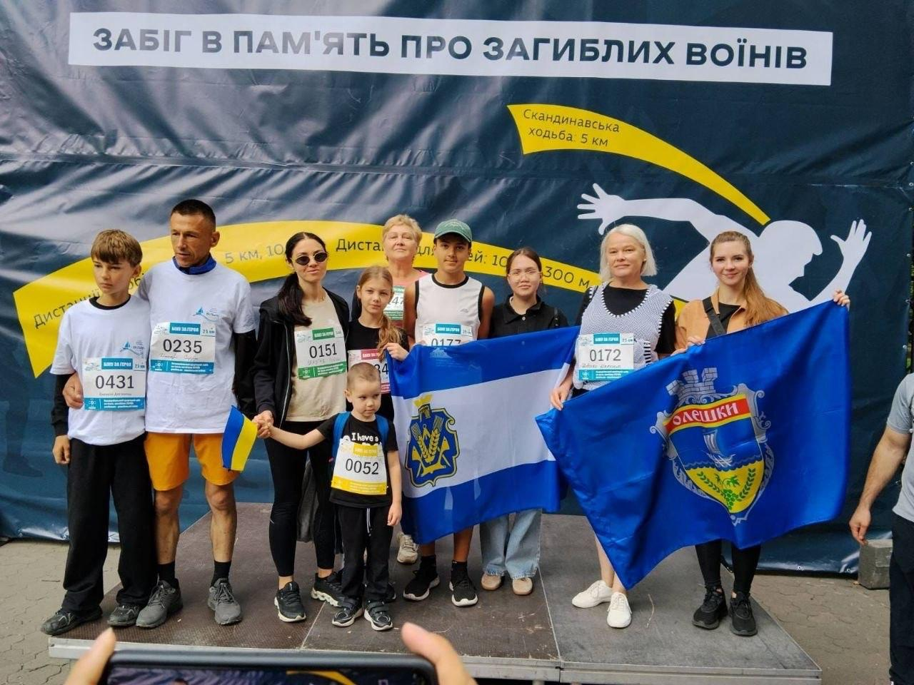 Дорослі та юні переселенці з Херсонщини — підопічні координаційного центру «Вільні разом» у Львові — позують на сцені після участі у Всеукраїнському забігу «Шаную воїнів, біжу за Героїв України». Група з десяти учасників у спортивному одязі та зі стартовими номерами забігу на грудях стоїть на тлі великого темно-синього банера з написом «ЗАБІГ В ПАМ'ЯТЬ ПРО ЗАГИБЛИХ ВОЇНІВ». Учасники у центрі та праворуч тримають два великі розгорнуті прапори: офіційний синьо-білий прапор Херсонської області з гербом регіону біля древка та синій прапор міста Олешки. Чоловік ліворуч тримає в руці маленький настільний прапорець України, а найменший учасник забігу попереду стоїть у фірмовій футболці з написом «I have a dream» (Я маю мрію).