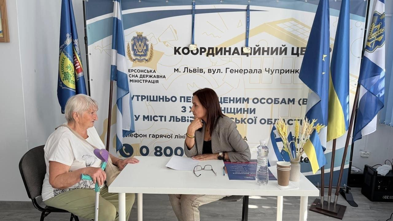 Начальниця Олешківської міської військової адміністрації Тетяна Гасаненко проводить особистий прийом громадян у координаційному центрі «Вільні разом» у місті Львів. Очільниця МВА у сірому піджаку в клітинку сидить за білим столом навпроти літньої жінки-переселенки, яка тримає в руці медичну підлікткову милицю з фіолетовою ручкою. На столі перед ними розкладено папери, ручку, окуляри та пляшку води. Поруч стоїть декоративний букет із колосків пшениці з маленьким прапорцем України. Зустріч проходить на тлі великого інформаційного банера Координаційного центру Херсонської ОВА.