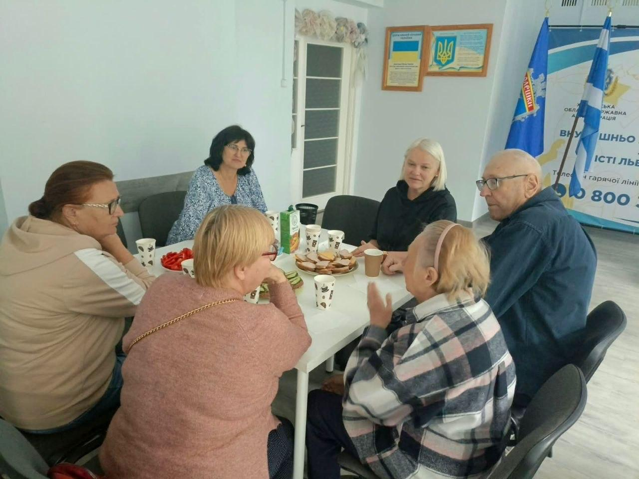 Група з шести переселенців з Херсонщини — п'ять жінок та літній чоловік в окулярах — сидять колом за білим столом у координаційному центрі «Вільні разом» у Львові під час психотерапевтичного заходу «Тепло своє передаю…». Учасники зустрічі п'ють чай, пригощаються солодощами, фруктами, бутербродами та ведуть довірливу бесіду у теплій атмосфері. У кінці столу сидить психологиня чи модераторка зустрічі у синій візерунчастій блузі, яка спрямовує хід терапії. Спілкування проходить на тлі великого офіційного банера Координаційного центру Херсонської ОВА.