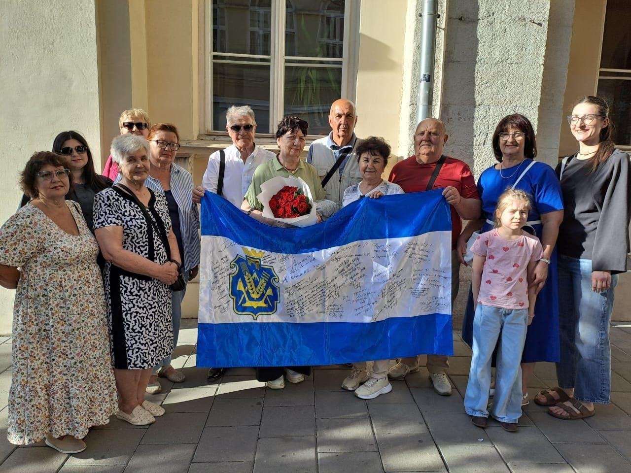 Група з дванадцяти дорослих переселенців з Херсонщини та одна маленька дівчинка позують для спільного фото на вулиці біля будівлі координаційного центру «Вільні разом» у Львові. Учасники зустрічі тримають перед собою великий розгорнутий офіційний прапор Херсонської області, біла смуга якого повністю вкрита рукописними побажаннями, словами підтримки та підписами земляків, зробленими маркером. Жінка в центрі тримає великий святковий букет червоних троянд у білій обгортці. Люди посміхаються та стоять на світлому тротуарі на тлі кам'яної стіни будівлі.