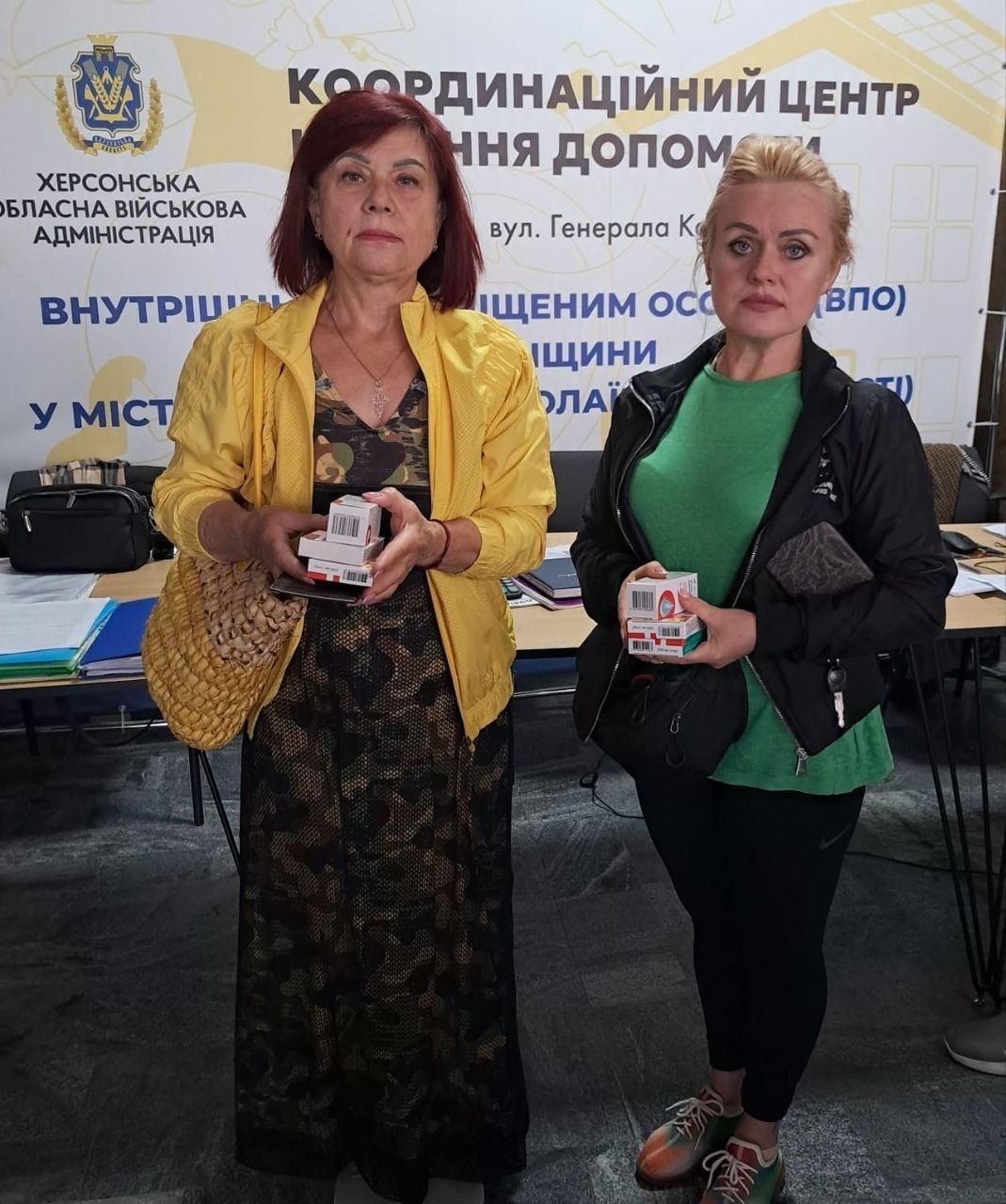 Дві жінки-переселенки з Херсонщини тримають у руках невеликі коробки з ліками та медичними виробами, отриманими від благодійного фонду «Медицина Херсонщини». Видача допомоги проходить у приміщенні координаційного центру «Вільні разом» у місті Миколаїв. Ліворуч стоїть жінка в камуфляжній сукні та яскравій жовтій куртці, праворуч — жінка в зеленій футболці та чорній спортивній кофті. Вони позують на тлі великого світлого банера Херсонської обласної військової адміністрації з офіційним гербом області та написами «КООРДИНАЦІЙНИЙ ЦЕНТР» і «ВНУТРІШНЬО ПЕРЕМІЩЕНИМ ОСОБАМ (ВПО) ХЕРСОНЩИНИ».