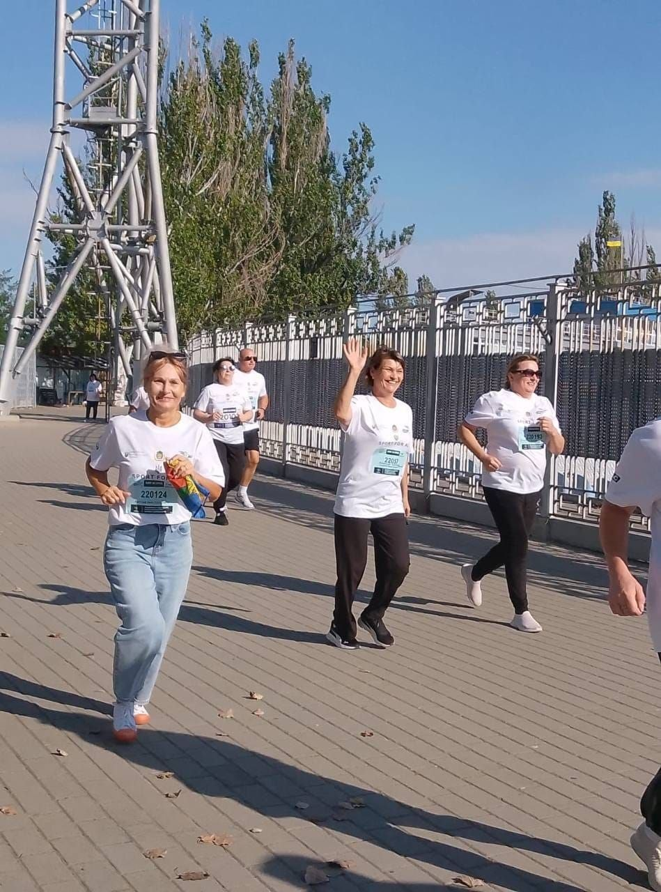 Переселенці з Херсонщини та фахівці координаційного центру «Вільні разом» у Миколаєві беруть участь у щорічному Всеукраїнському патріотичному забігу «Шаную воїнів, біжу за Героїв України». Учасники у сонцезахисних окулярах та однакових білих фірмових футболках зі стартовими номерами біжать дистанцію по брукованій доріжці в сонячний день. Попереду ліворуч біжить усміхнена жінка в блакитних джинсах зі стартовим номером 220124, яка тримає в руці невеликий згорнутий прапор України. Трохи позаду по центру біжить інша учасниця у чорних спортивних штанях зі стартовим номером 220017, яка посміхається та вітально махає рукою в камеру. Захід проходить уздовж довгого металевого паркану на тлі великої залізної конструкції та зелених дерев під яскравим синім небом.