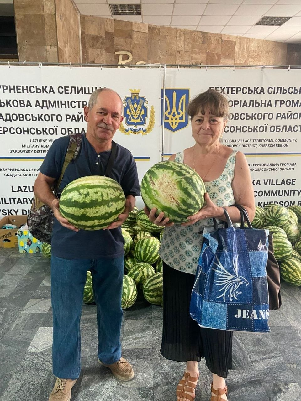 Чоловік і жінка літнього віку — переселенці з Херсонщини — посміхаються та тримають у руках по великому стиглому смугастому кавуну, отриманому від фермера Володимира Галичиновського. Видача баштанних проходить у приміщенні координаційного центру «Вільні разом» у місті Миколаїв. Чоловік ліворуч одягнений у темно-синю футболку та джинси, жінка праворуч — у світлу літню блузу та чорну спідницю, у руці вона тримає велику синю сумку. Вони позують на тлі білого банера Лазурненської селищної військової адміністрації та Бехтерської сільської громади Скадовського району із зображенням державних гербів. Позаду людей на підлозі складено велику купу інших кавунів.