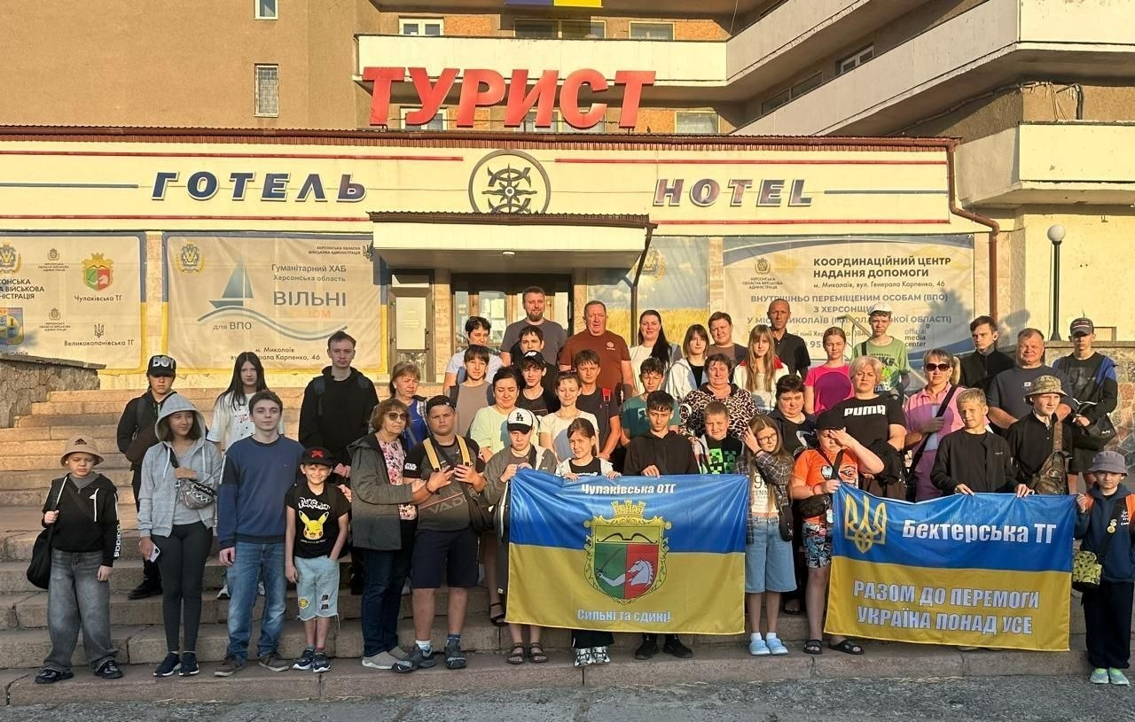 Велика група дорослих, підлітків та дітей-переселенців із Херсонщини позує для спільного фото на сходах перед евакуаційним виїздом на відпочинок. Збір учасників проходить біля входу до готелю «Турист» у місті Миколаїв, де працює координаційний центр «Вільні разом». Попереду діти та батьки тримають два великі розгорнуті прапори своїх рідних куточків Херсонщини: жовто-блакитний прапор Чулаківської ОТГ із гербом і гаслом «Сильні та єдині!» та синьо-жовтий прапор Бехтерської територіальної громади з написом «Разом до перемоги. Україна понад усе». Позаду групи на фасаді будівлі розміщено офіційні банери гуманітарного хабу та координаційного центру надання допомоги ВПО.
