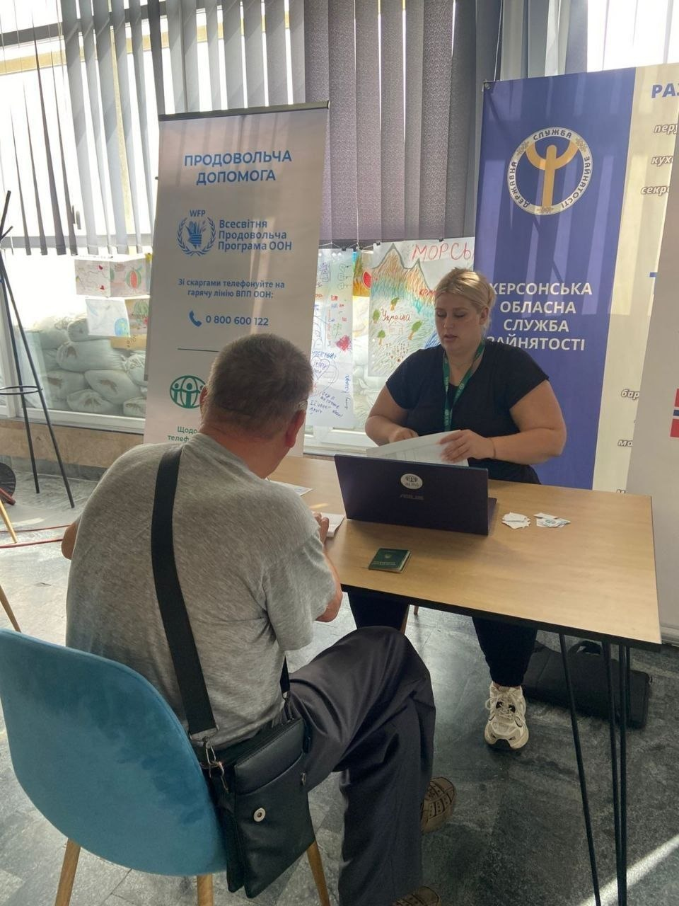 Індивідуальна юридична консультація переселенця з Херсонщини в координаційному центрі «Вільні разом» у місті Миколаїв. На передньому плані спиною до камери на синьому стільці сидить чоловік у сірій футболці із сумкою через плече, на столі перед ним лежить паспорт у зеленій обкладинці. Навпроти нього сидить юрисконсультантка — світловолоса жінка у чорній футболці, яка працює за відкритим ноутбуком та вивчає паперові документи. Праворуч за нею встановлено темно-синій банер із написом «ХЕРСОНСЬКА ОБЛАСНА СЛУЖБА ЗАЙНЯТОСТІ», а ліворуч — білий вертикальний ролап із написом «ПРОДОВОЛЬЧА ДОПОМОГА / Всесвітня продовольча програма ООН (WFP)».