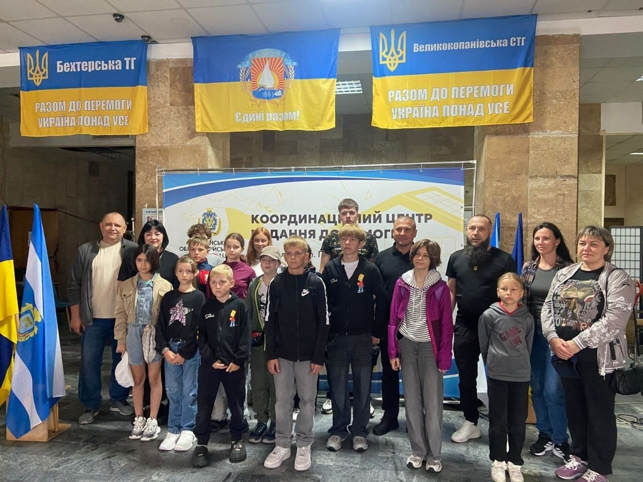 Група переселенців із Херсонщини різного віку позує для спільного фото всередині миколаївського координаційного центру «Вільні разом». Крайній ліворуч стоїть начальник Великокопанівської СВА Олексій Соловйов у світлій футболці та темній сорочці. Учасники зустрічі розташовані перед великим білим банером «КООРДИНАЦІЙНИЙ ЦЕНТР НАДАННЯ ДОПОМОГИ». На стіні над ними розвішано три великі синьо-жовті прапори: ліворуч — Бехтерської ТГ, по центру — прапор із написом «Єдині разом!», праворуч — Великокопанівської СТГ. На стягах Бехтерської та Великокопанівської громад нанесено гасло «РАЗОМ ДО ПЕРЕМОГИ. УКРАЇНА ПОНАД УСЕ». 