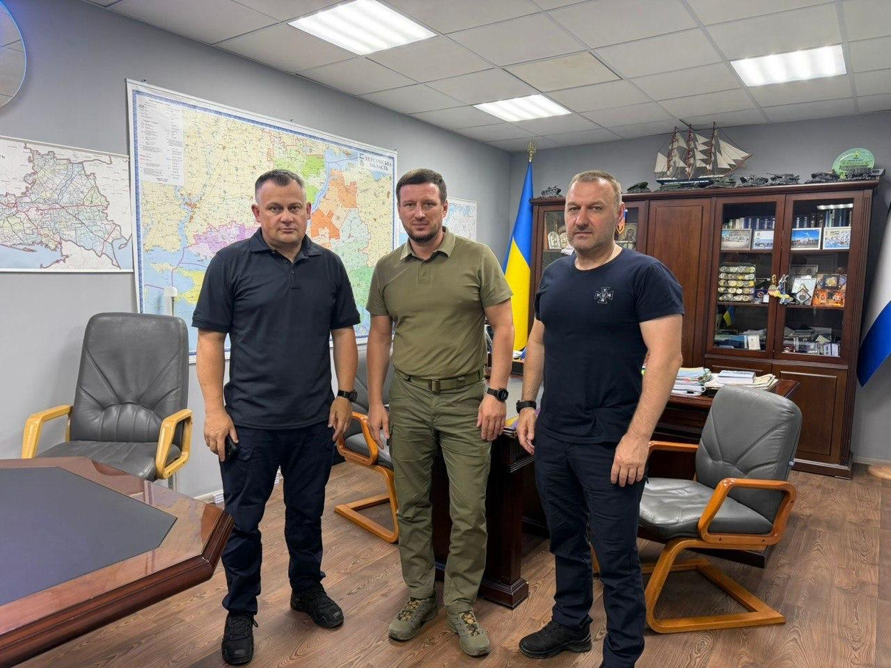 Начальник Херсонської ОВА Олександр Прокудін, Голова ДСНС України Андрій Даник та його Перший заступник Віталій Миронюк стоять разом у ряд по центру робочого кабінету під час офіційної зустрічі у Херсоні. По центру стоїть Олександр Прокудін у тактичній футболці та штанах кольору хакі (олива). Ліворуч від нього стоїть Голова ДСНС Андрій Даник у чорній футболці-поло та чорних тактичних штанах. Праворуч — Перший заступник Голови ДСНС Віталій Миронюк у чорній футболці з невеликим емблемним Тризубом на грудях та чорних штанах. Чоловіки позують біля великого масивного дерев'яного столу засідань. На сірій стіні позаду висять детальні карти Херсонської області та України, а в кутку кімнати встановлено державний прапор України та дерев'яну книжкову шафу.