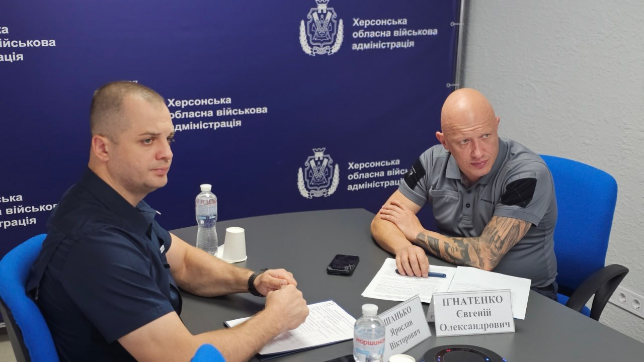 Робоча нарада щодо евакуації родин із дітьми на Херсонщині. Заступник начальника Херсонської ОВА Євгеній Ігнатенко (праворуч) та начальник Херсонської МВА Ярослав Шанько (ліворуч) сидять за столом у залі засідань на тлі синього банера з офіційною символікою «Херсонська обласна військова адміністрація». Посадовці координують безпечне вивезення та створення умов для проживання родин із дітьми, які досі залишаються в небезпечних зонах деокупованої частини Херсонщини.