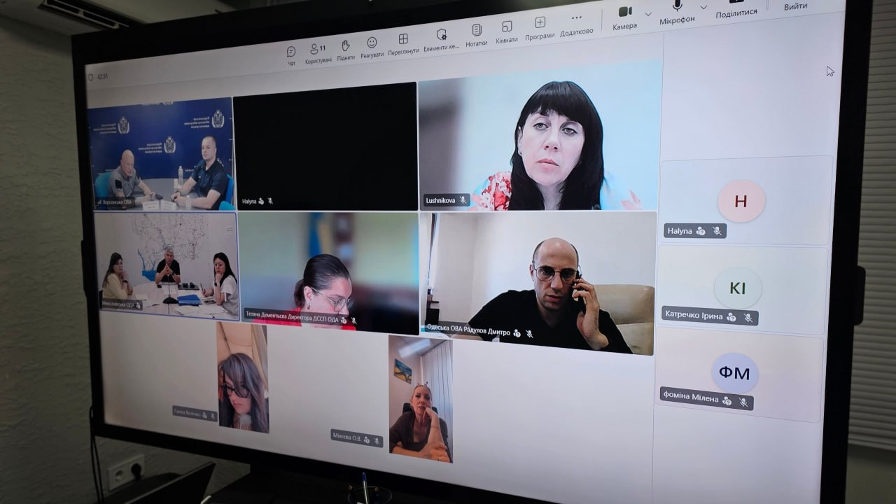 Екран монітора з онлайн-нарадою у програмі Microsoft Teams щодо евакуації родин із дітьми на Херсонщині. На великому дисплеї відображається сітка з відеовікнами учасників зустрічі. В окремих вікнах присутні: у верхньому лівому кутку — керівництво у залі засідань Херсонської ОВА (заступник начальника ОВА Євгеній Ігнатенко та начальник Херсонської МВА Ярослав Шанько); поруч — вікно з підписом «Lushnikova», де присутня начальниця служби у справах дітей Херсонської ОВА Тетяна Лушнікова; нижче — директорка Департаменту соціальної та сімейної політики Одеської ОДА Тетяна Дементьєва, а також заступник голови Одеської ОВА Дмитро Радулов (спілкується по телефону). Справа виведено список інших учасників наради без увімкнених камер.