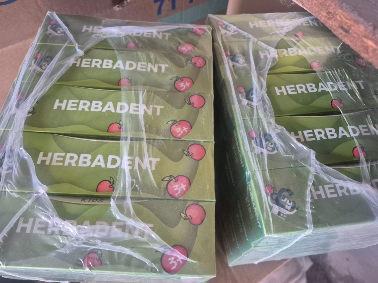 Упаковки дитячої зубної пасти бренду Herbadent, що надійшли як гуманітарна допомога від організації «Шанс на нове життя» для сімей у модульному містечку на Херсонщині. На фотографії зблизька зображено дві великі пачки, загорнуті у прозову пакувальну плівку, всередині яких щільно складені зелені картонні коробки із дитячою зубною пастою «Herbadent Kids». На кожній коробці міститься малюнок червоних яблук та рожеве маркування «3+», що вказує на вікову категорію засобів гігієни для дітей від трьох років. Партія стоматологічних наборів зафіксована у процесі розвантаження та передачі юним жителям області.