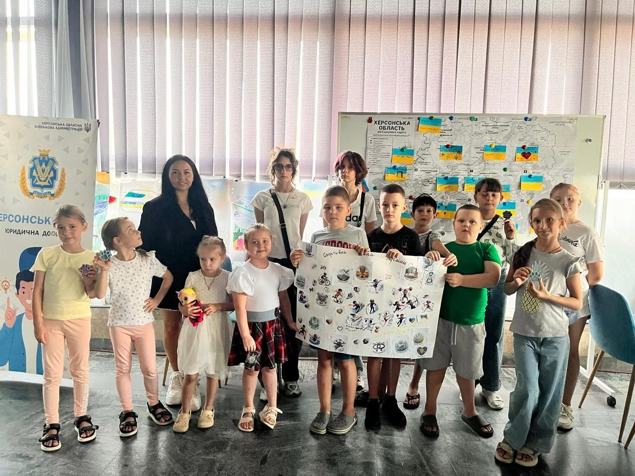 Група з тринадцяти дітей-переселенців із Херсонщини та одна жінка-організаторка у чорному піджаку (стоїть ліворуч на передньому плані) позують для спільного фото в миколаївському координаційному центрі «Вільні разом». Після завершення артзаходу хлопчик у центрі тримає великий створений дітьми плакат-колаж під назвою «Спортивна мапа України», вкритий наліпками з різними видами спорту. Інші діти дошкільного та шкільного віку з посмішками тримають у руках власноруч виготовлені яскраві брелоки-шейкери у формі лапок та патріотичних сердечок. На задньому плані стоїть мобільний стенд із детальною інтерактивною картою «Херсонська область», на якій населені пункти прикрашені маленькими паперовими прапорцями України. Ліворуч видно вертикальний ролап із гербом Херсонської ОВА та написом «Юридична допомога».