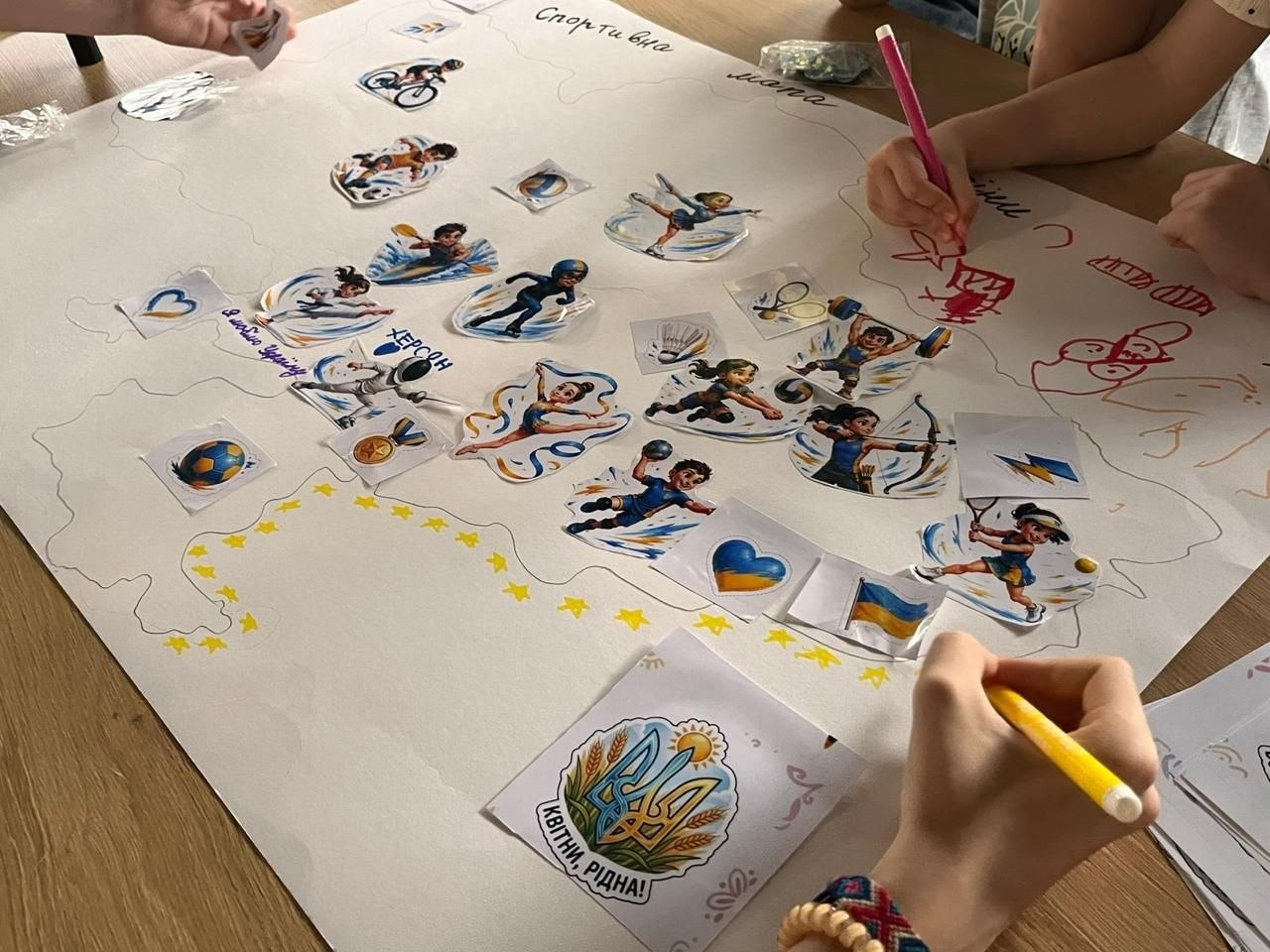 Крупний план великого паперового ватмана з контуром карти України під час інтерактивного дитячого заняття «Спортивна мапа України» в миколаївському координаційному центрі «Вільні разом». У верхній частині карти від руки нанесено назву «Спортивна мапа». По всій її території діти-переселенці з Херсонщини розклеїли яскраві наліпки із зображеннями різних видів спорту: велоспорту, плаванням, художньою гімнастикою, футболом, тенісом та стрільбою з лука. На території Херсонської області нанесено великий рукописний напис синім маркером «ХЕРСОН», а на південних кордонах наклеєно лінію з маленьких жовтих зірочок, сердець та прапорців України. У нижній частині колажу прикріплено малюнок із гербом-Тризубом, соняшником та гаслом «Квітни, рідна!». Навколо плаката видно дитячі руки з кольоровими фломастерами, які домальовують контури та візерунки.