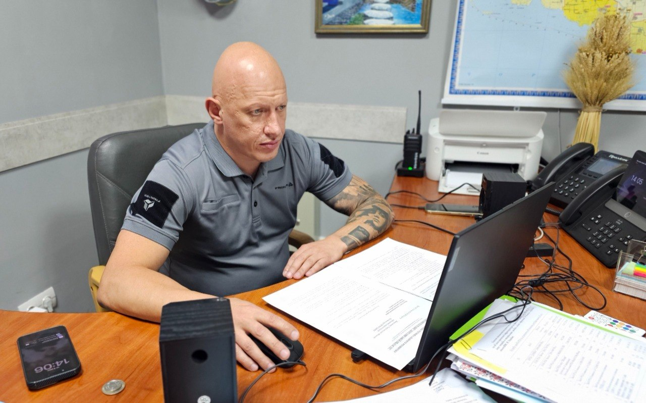 Заступник начальника Херсонської обласної військової адміністрації Євгеній Ігнатенко працює за робочим столом у службовому кабінеті під час онлайн-зустрічі щодо аналізу роботи партнерських організацій у медичній сфері та визначення потреб лікарень і амбулаторій Бериславського району. Посадовець одягнений у сіру сорочку-поло з нашивками на рукавах. Він зосереджено дивиться на екран відкритого чорного ноутбука, тримаючи праву руку на комп'ютерній миші, а ліву — на роздрукованих паперових документах. 