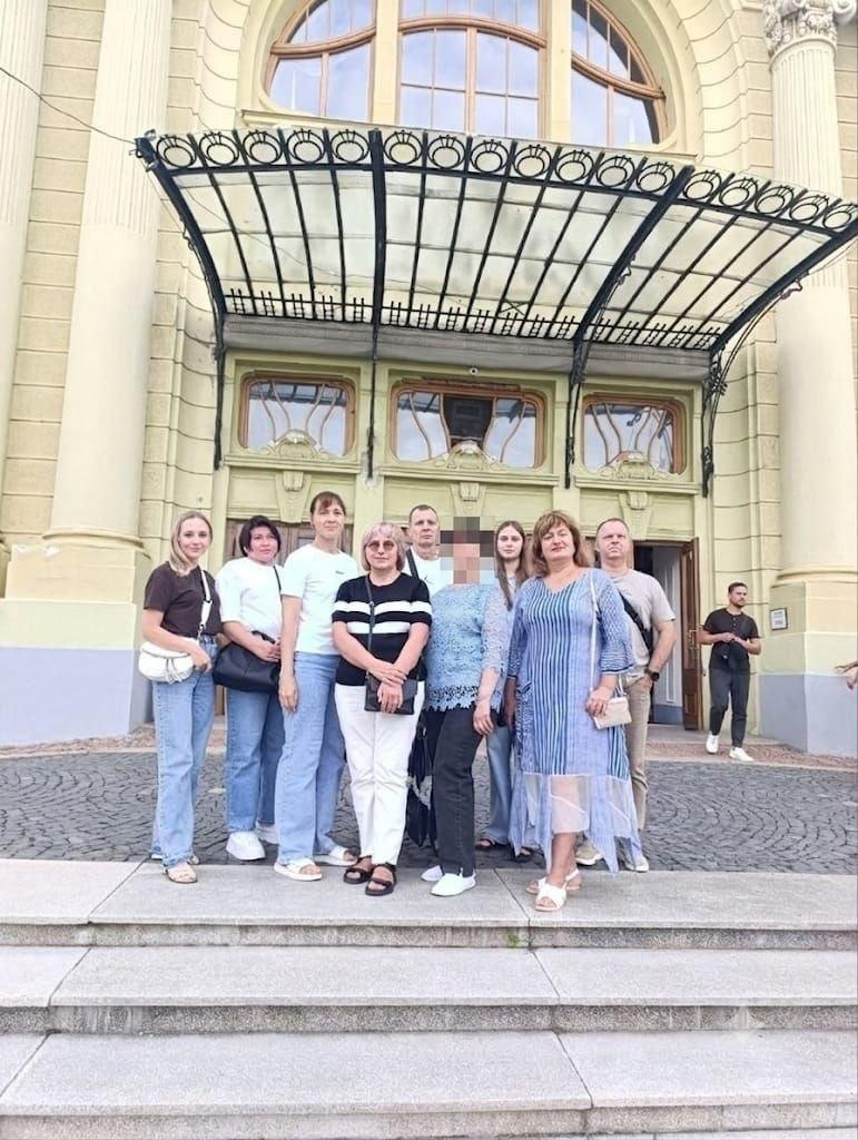 Група переселенців із Херсонщини — сім жінок та два чоловіки — позують для спільного фото на кам'яних сходах перед головним входом до Чернівецького академічного обласного українського музично-драматичного театру імені Ольги Кобилянської. Обличчя однієї з жінок у центрі кадру розмите цифровим способом (затушоване). Візит організовано координаційним центром «Вільні разом» у Чернівцях, де ВПО стали глядачами вистави «Кайдашева сім'я». Учасники заходу посміхаються та стоять разом на тлі величного світло-жовтого фасаду історичної будівлі театру, прикрашеного масивними колонами та зеленими дерев'яними дверима. Над центральним входом безпосередньо над групою людей встановлено великий старовинний вигнутий козирок із чорного кованого металу та скла. На задньому плані праворуч на бруківці стоїть ще один перехожий чоловік у чорній футболці.