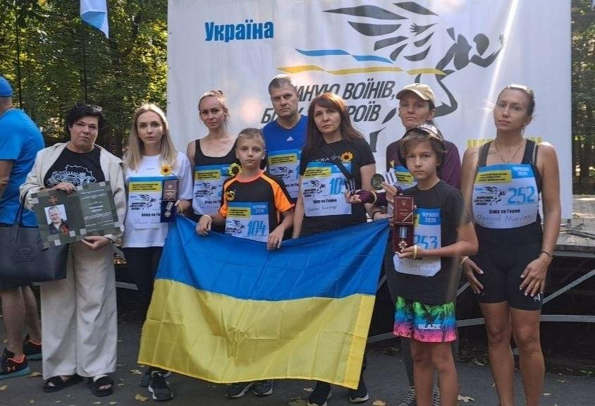 Колектив координаційного центру «Вільні разом» у Чернівцях та переселенці з Херсонщини позують для спільного фото під час участі у Всеукраїнському патріотичному забігу «Шаную воїнів. Біжу за Героїв України». Група з шести жінок, одного чоловіка та двох хлопчиків стоїть у спортивному одязі зі стартовими номерами на грудях (зокрема 104, 252, 253) перед великим світлим банером заходу, на якому написано «Україна» та «ШАНУЮ ВОЇНІВ, БІЖУ ЗА ГЕРОЇВ». Учасники тримають перед собою великий розгорнутий державний прапор України. Крайня ліворуч жінка тримає відкриту зелену папку-диплом із фотографією загиблого українського захисника, а дівчина у білій футболці поруч із нею та хлопчик на передньому плані тримають у руках державні нагороди й медалі у футлярах, біля яких прикріплено квіти соняшника. Крайнім ліворуч на задньому плані спиною до камери стоїть інший чоловік у синій футболці та синій кепці, який також тримає велику чорну господарську сумку.